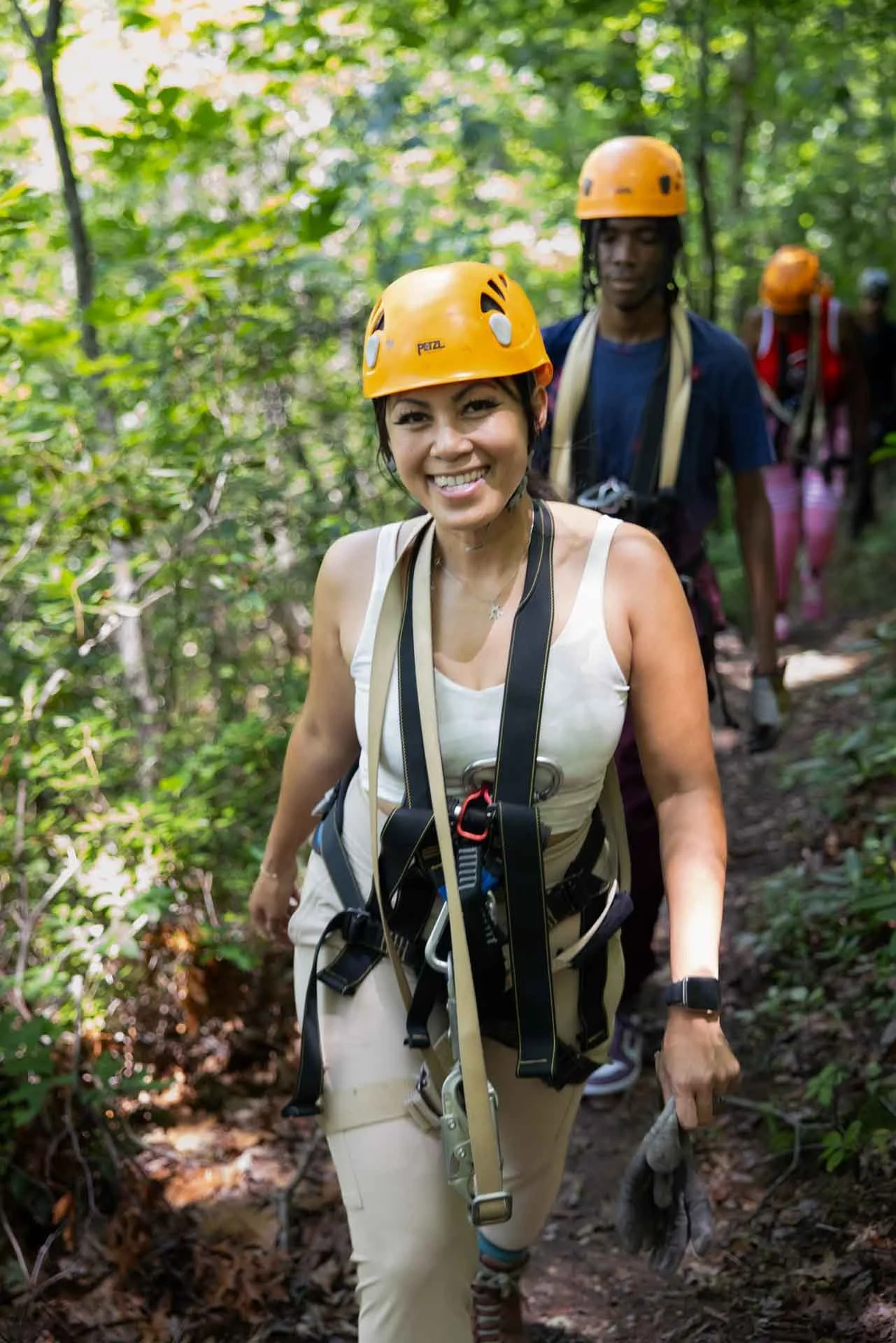 A woman smiling as she hikes through a forest wearing a yellow helmet and adventure gear. Behind her, other hikers wearing helmets follow along the trail.