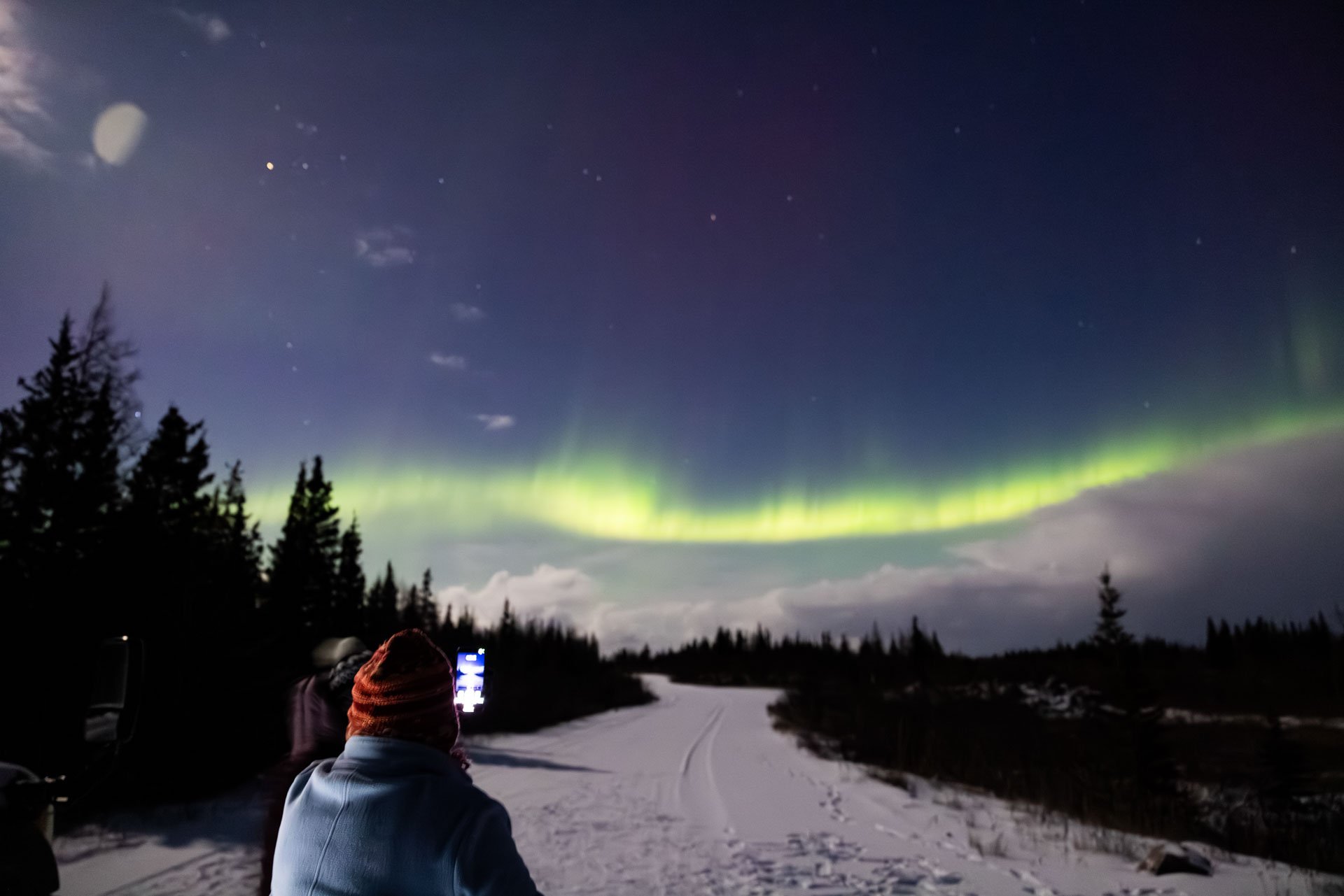 People viewing the northern lights in a snowy landscape with trees, night sky, and visible stars and clouds.