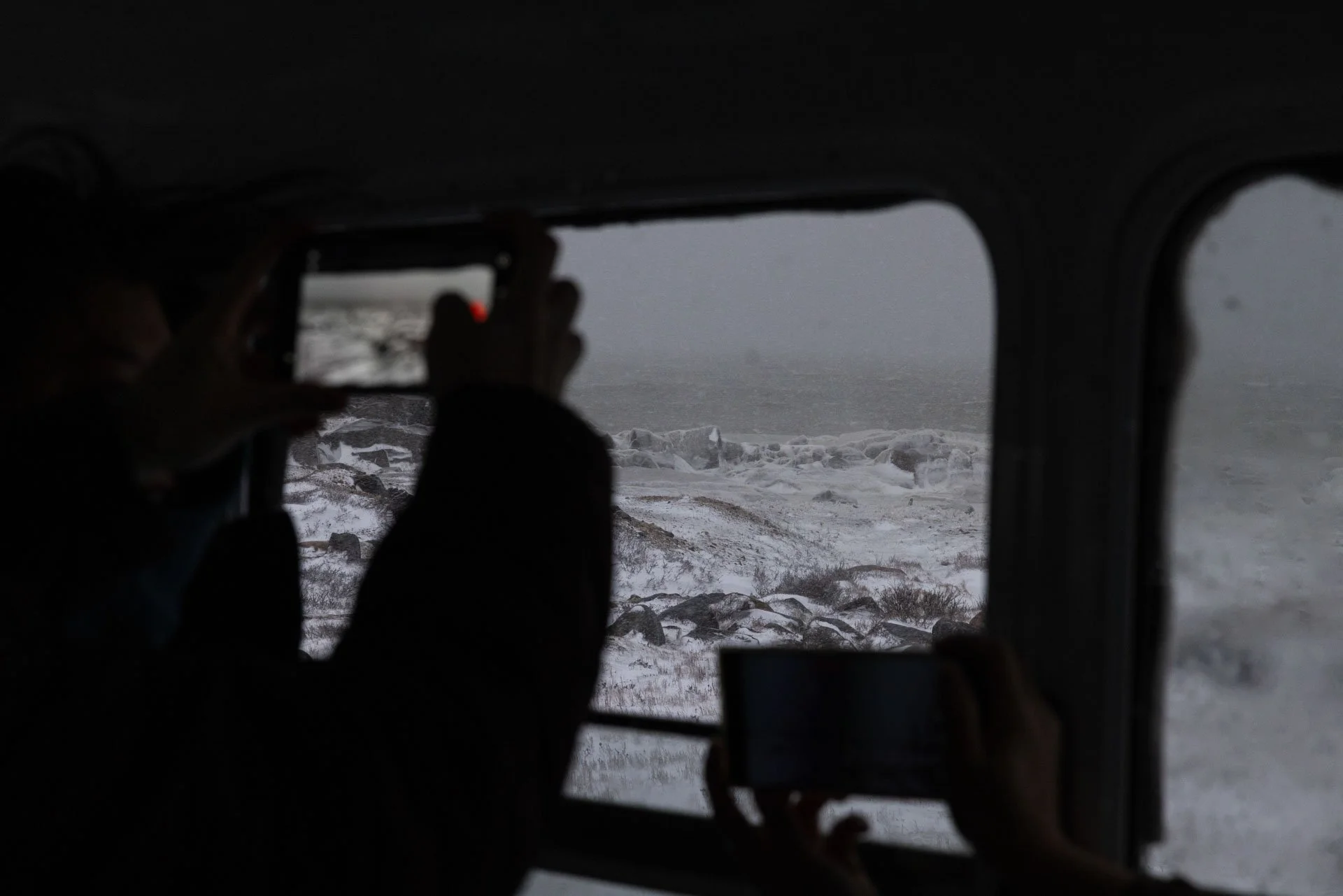 People inside a vehicle taking photos of a snowy, rocky shoreline and overcast sky through a window.