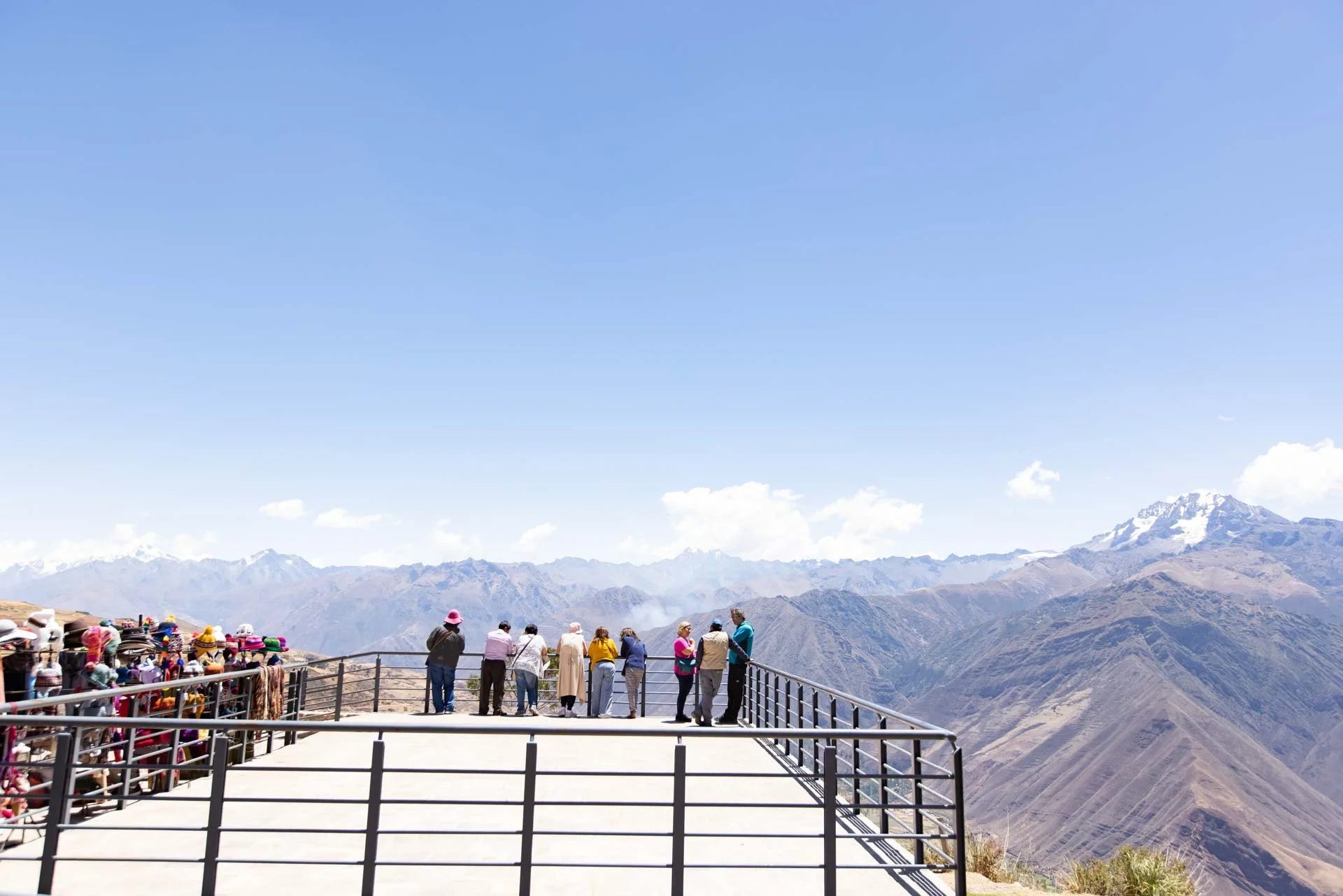 Tourists standing on a viewing platform overlooking mountains with snow-capped peaks in the distance under a clear blue sky, with some vendor stalls on the left.
