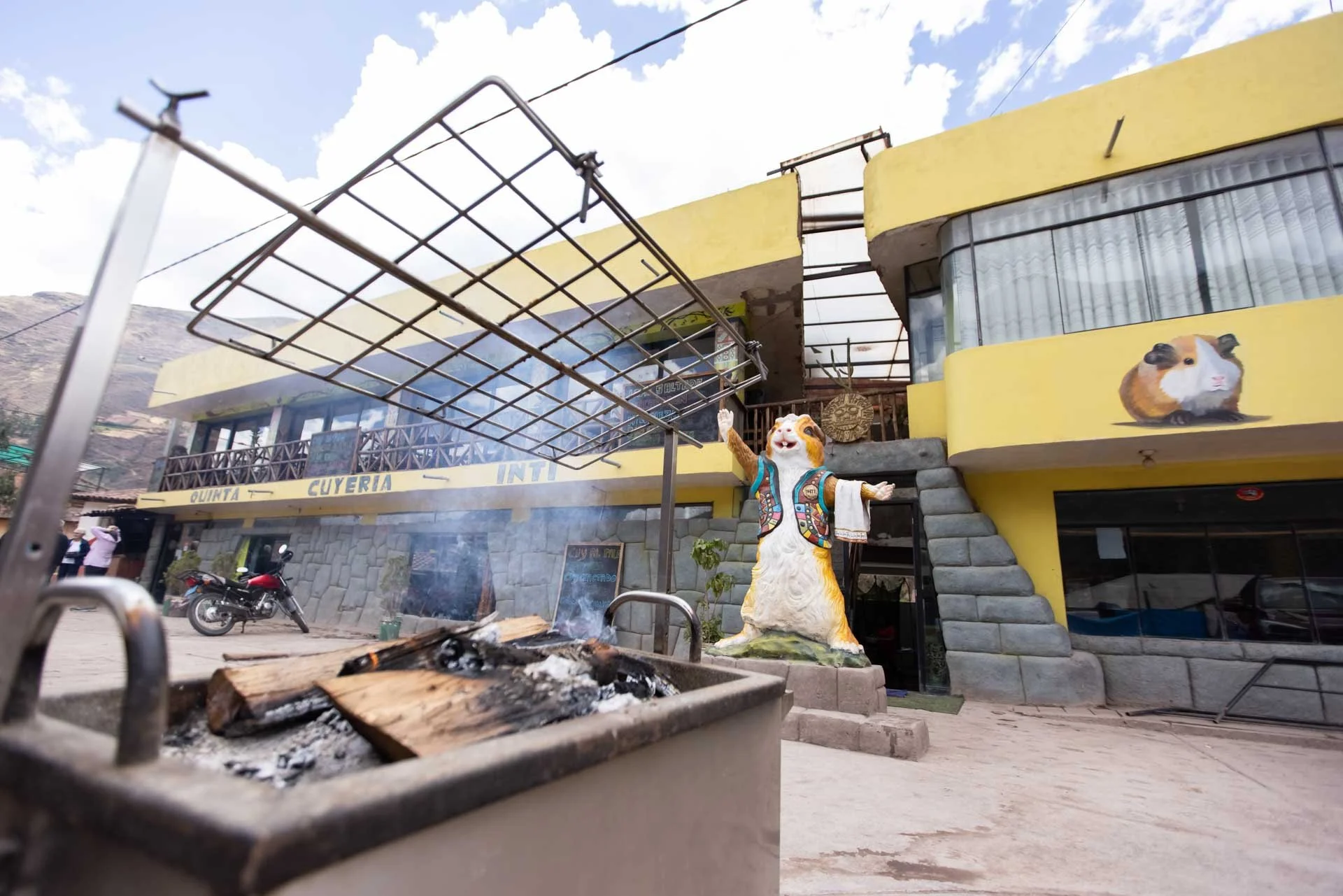 A building with a yellow facade and outdoor seating, decorated with guinea pig images on the walls. A statue of a guinea pig dressed in a jacket with arms outstretched is near the entrance. There is a small outdoor fireplace with burning wood and smo
