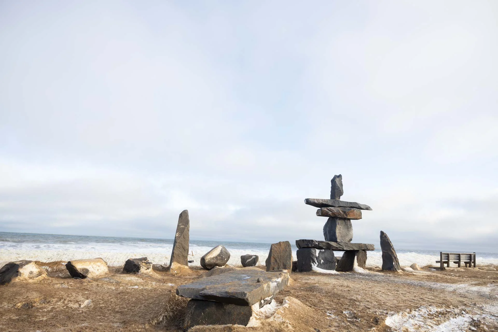 Beach with stone sculptures, a bench, and ocean waves in the background, under a partly cloudy sky.