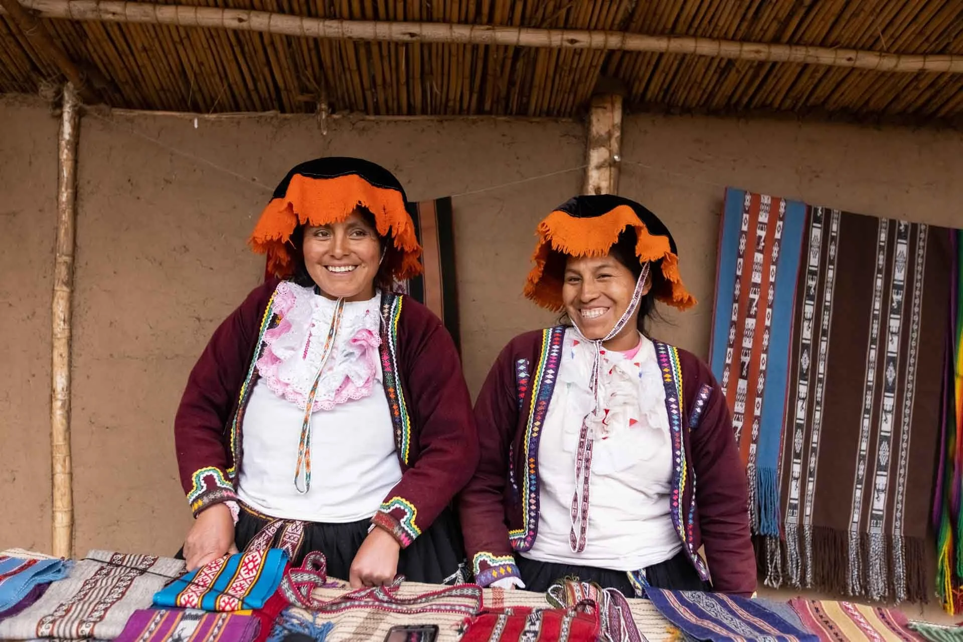Two women wearing traditional Andean clothing with orange and black hats, smiling at a market stall displaying colorful textiles and crafts.