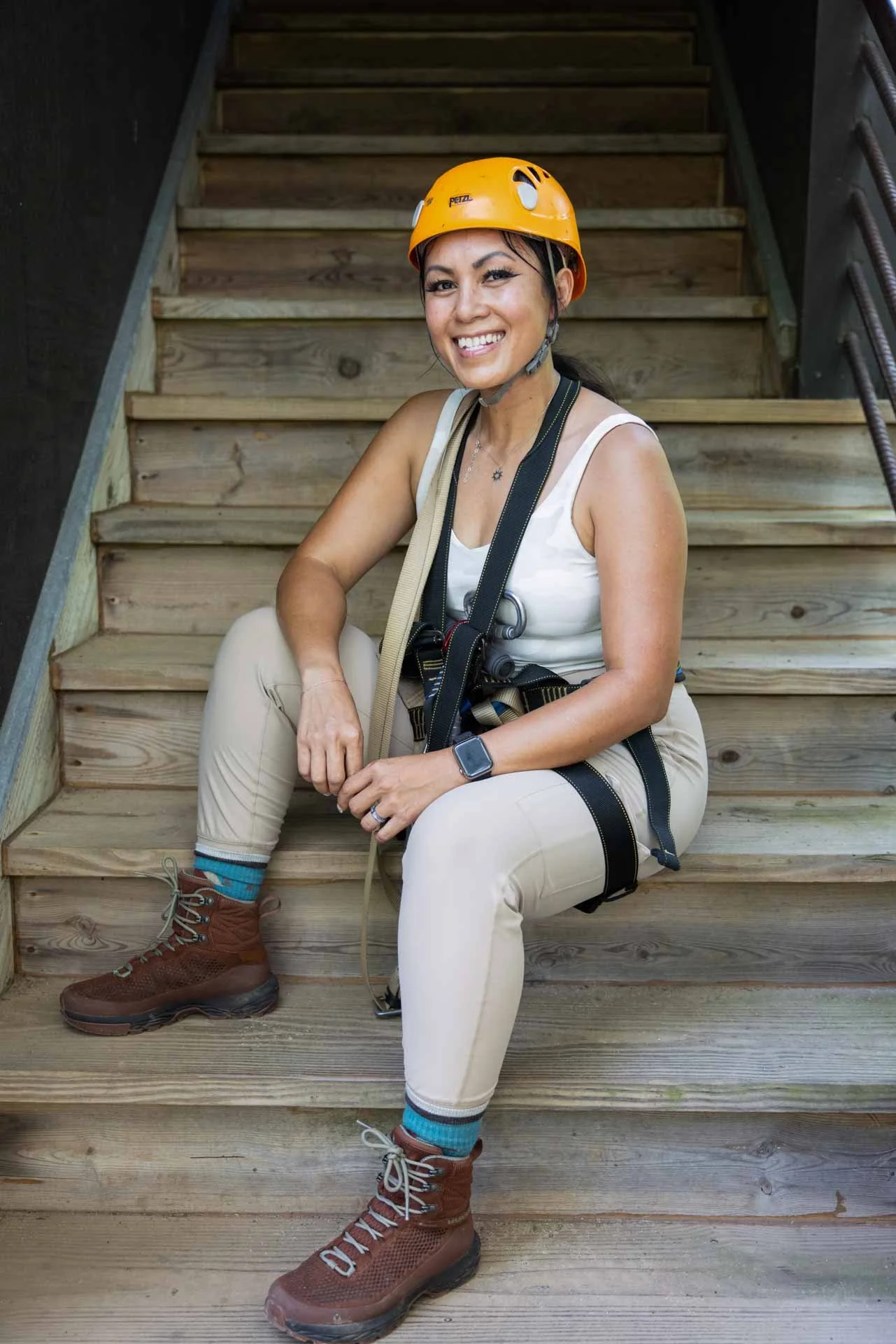 A woman in a white tank top, khaki pants, and brown hiking boots is sitting on wooden stairs, smiling, wearing an orange safety helmet, a watch, and a camera around her neck.