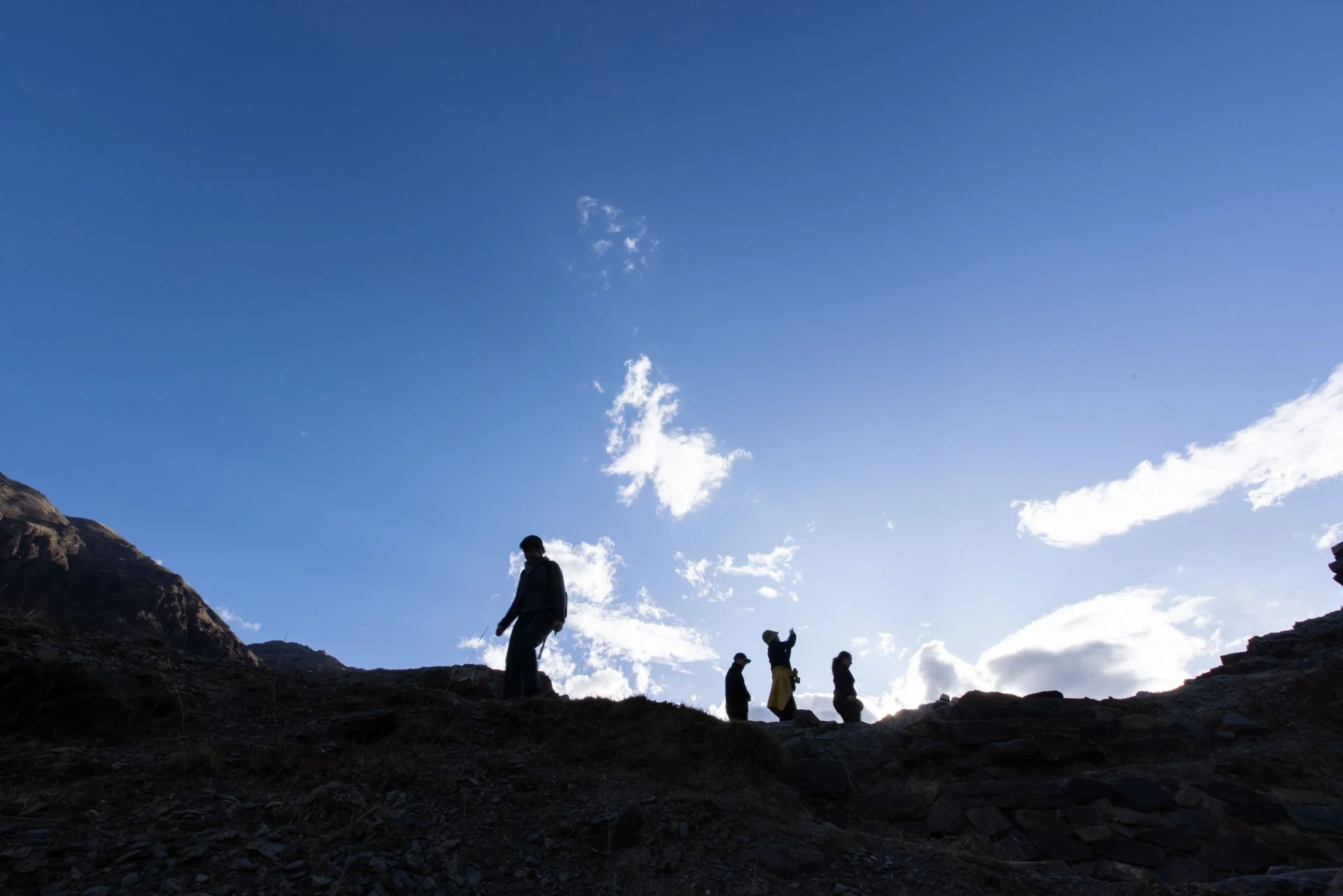 Four people walking along a rocky trail on a mountain under a bright blue sky with a few white clouds.
