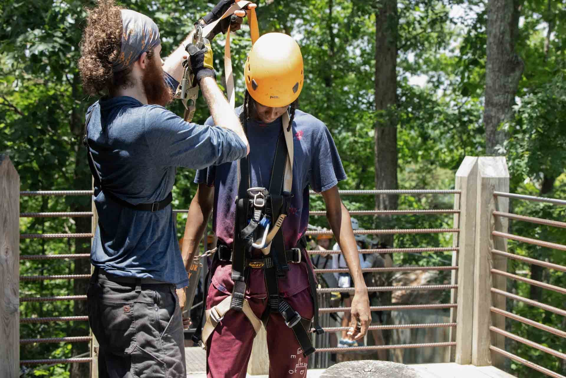Two men are outdoors on a wooden platform, with one adjusting the other's safety harness while the other wears a yellow helmet. There are trees and other people in the background.
