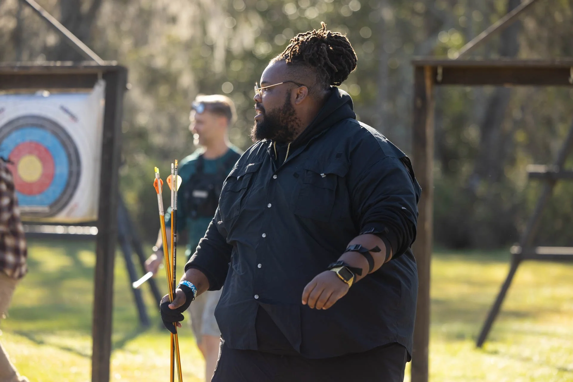 A man with dreadlocks and a beard holding a set of colorful arrows on an outdoor archery range, with a person in the background near an archery target.