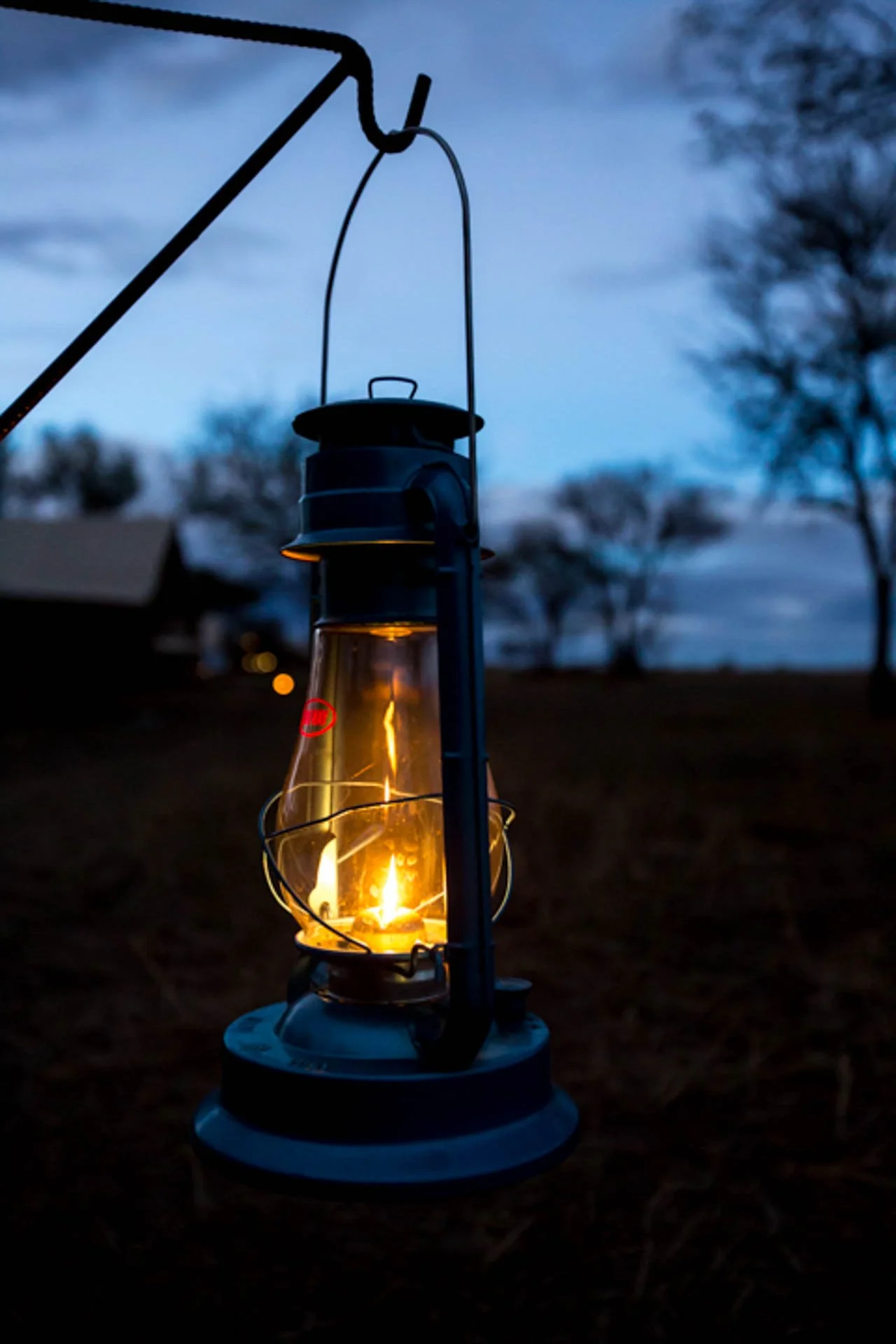 A lit lantern hanging outside during dusk with trees and a house visible in the background.