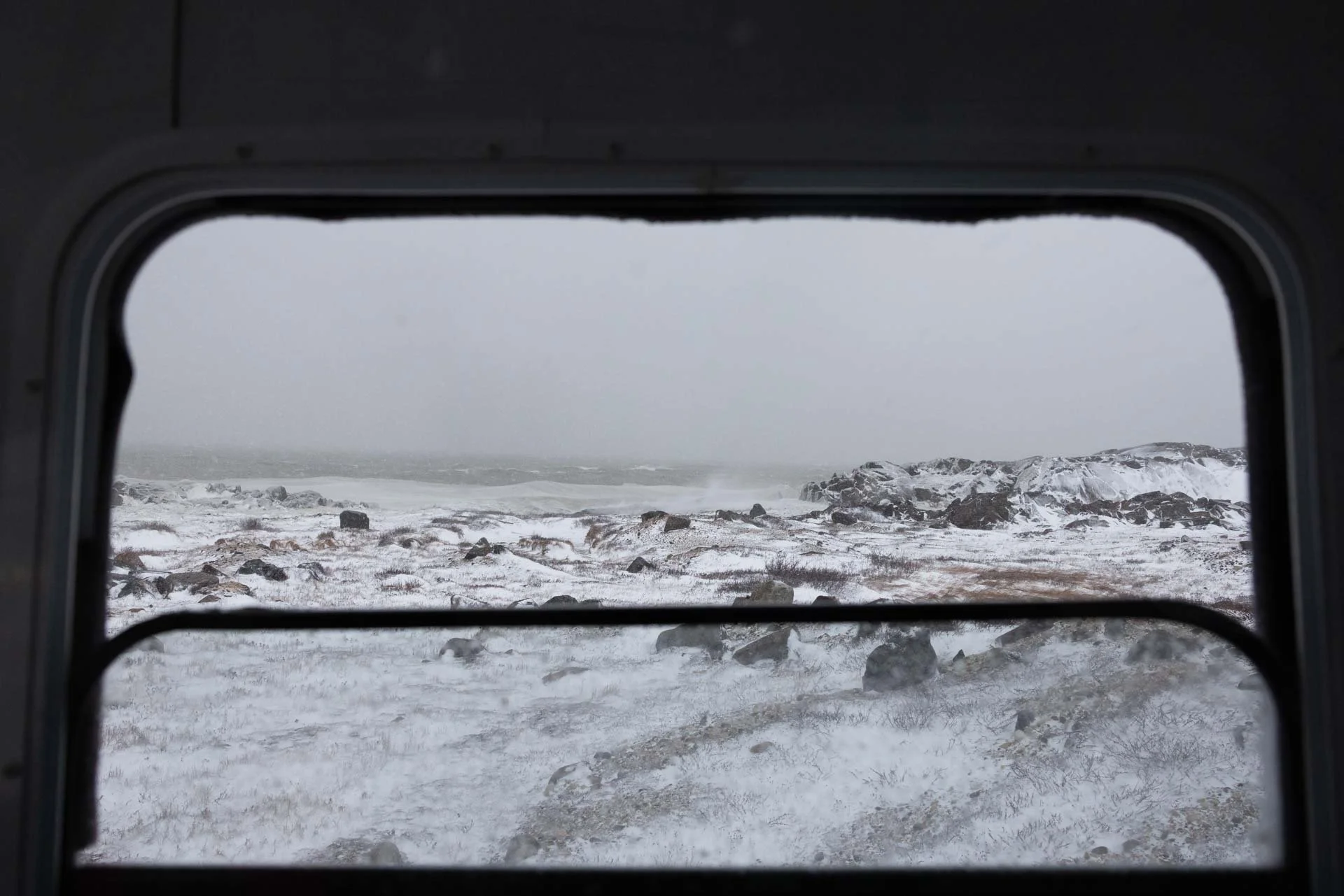 Snowy coastal landscape seen through a window, with rocky terrain and overcast skies.