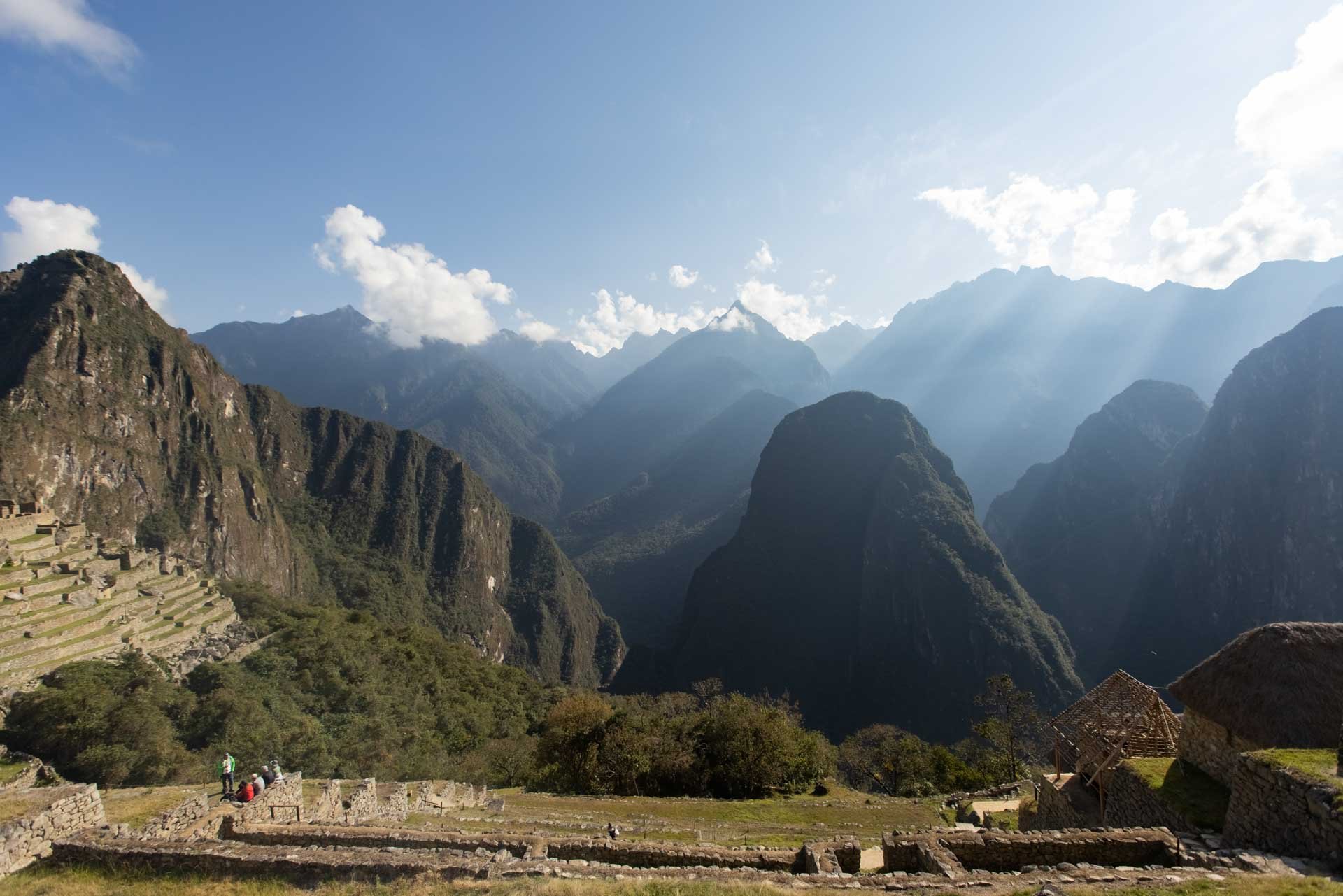 Ancient Incan ruins with terraced fields in the foreground, surrounded by lush green mountains and towering peaks under a partly cloudy sky.