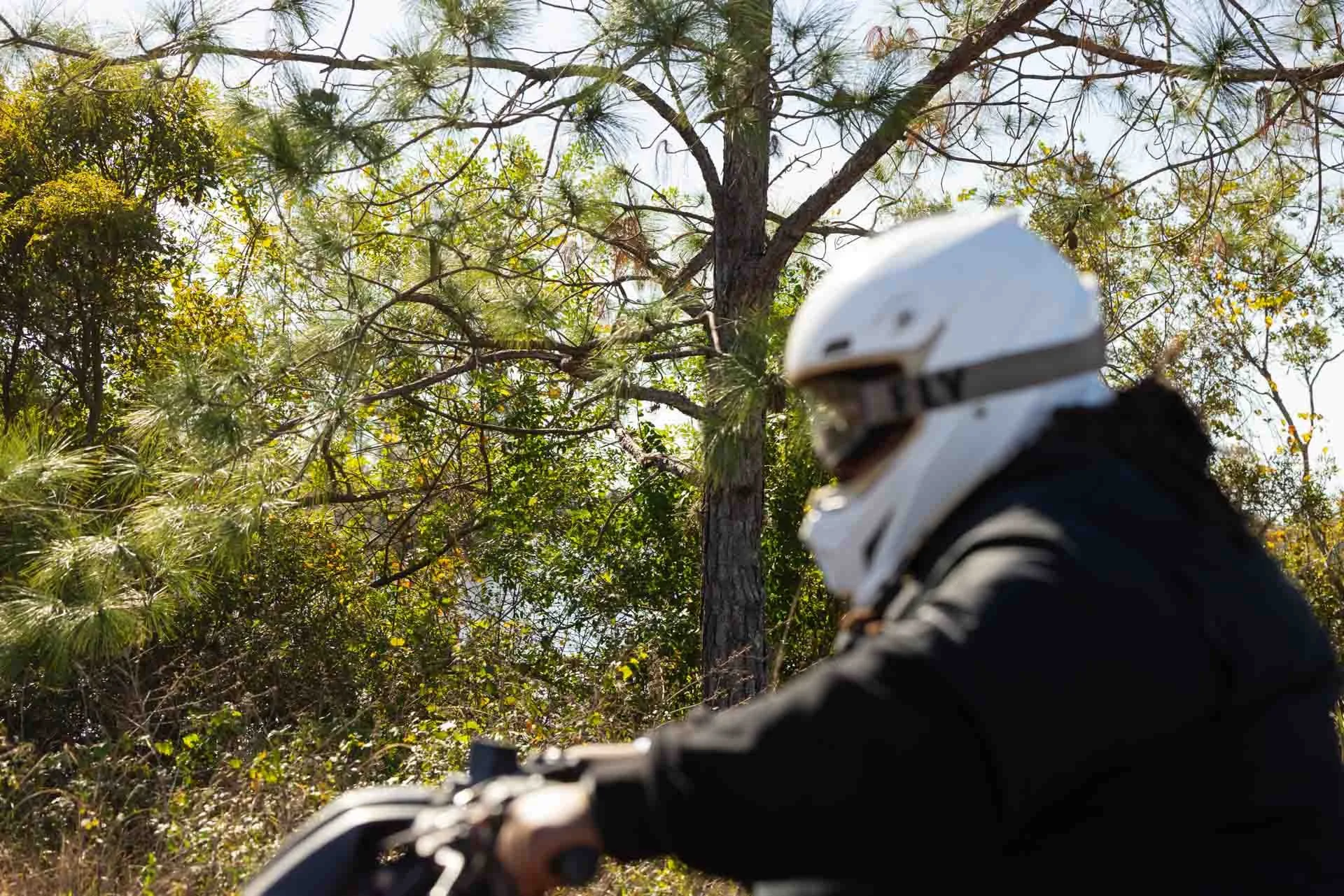 A person wearing a white helmet riding a motorcycle through a forested area with green trees and sunlight.