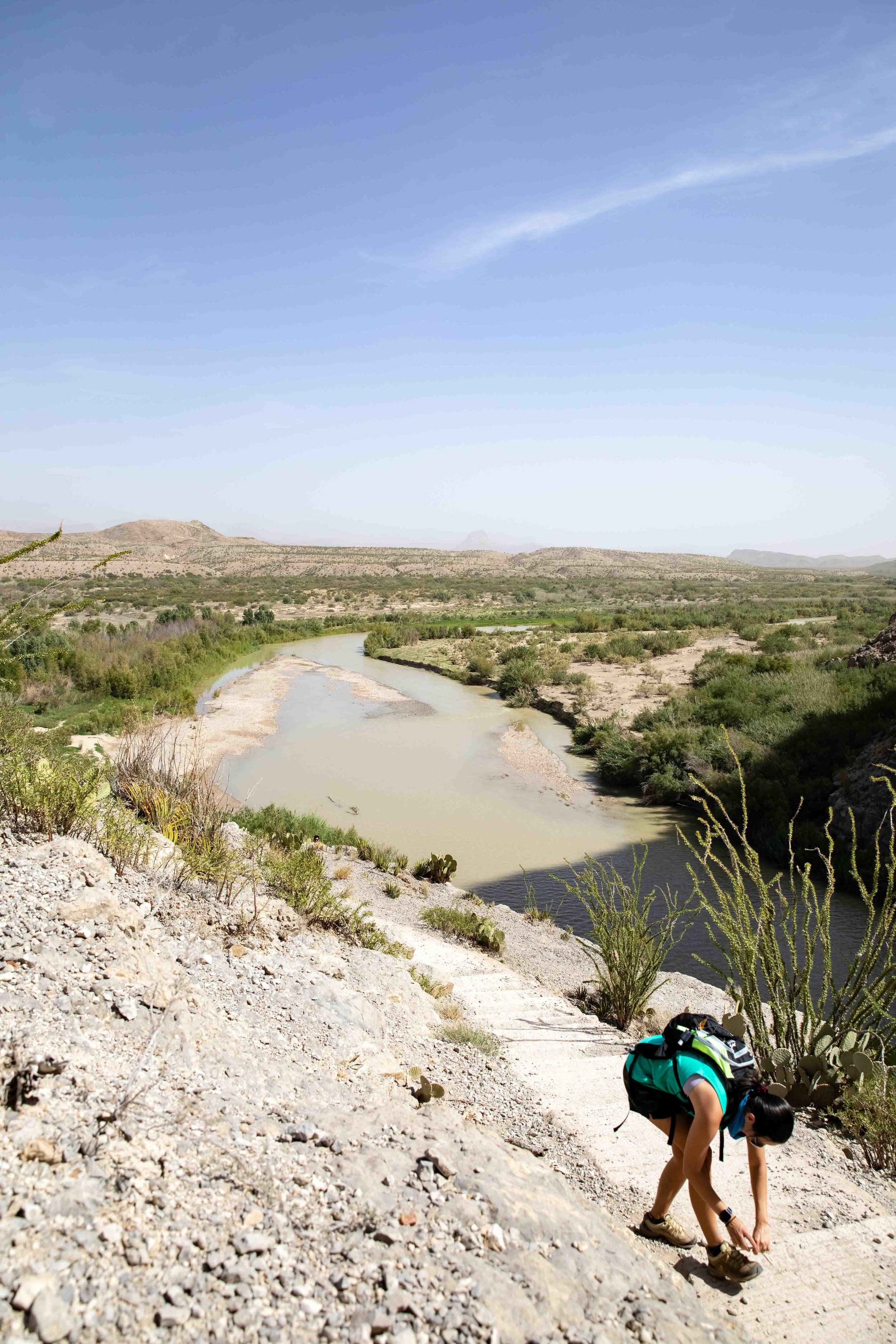 A person with a backpack tying their hiking shoelaces on a dirt trail near a river in a desert landscape, with hills and a clear blue sky in the background.