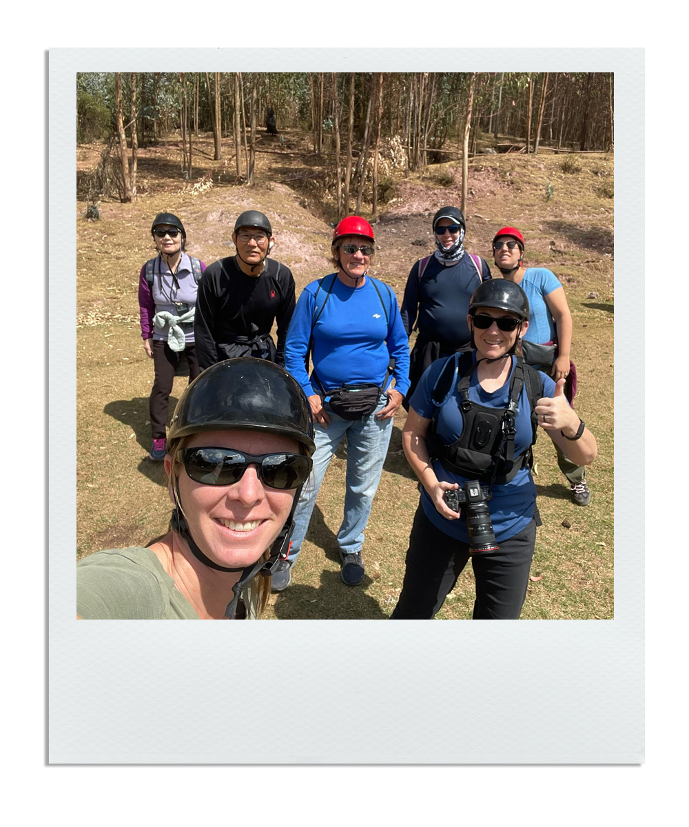 Group of seven people outdoors, wearing helmets and sunglasses, some with backpacks, taking a selfie in a wooded area.