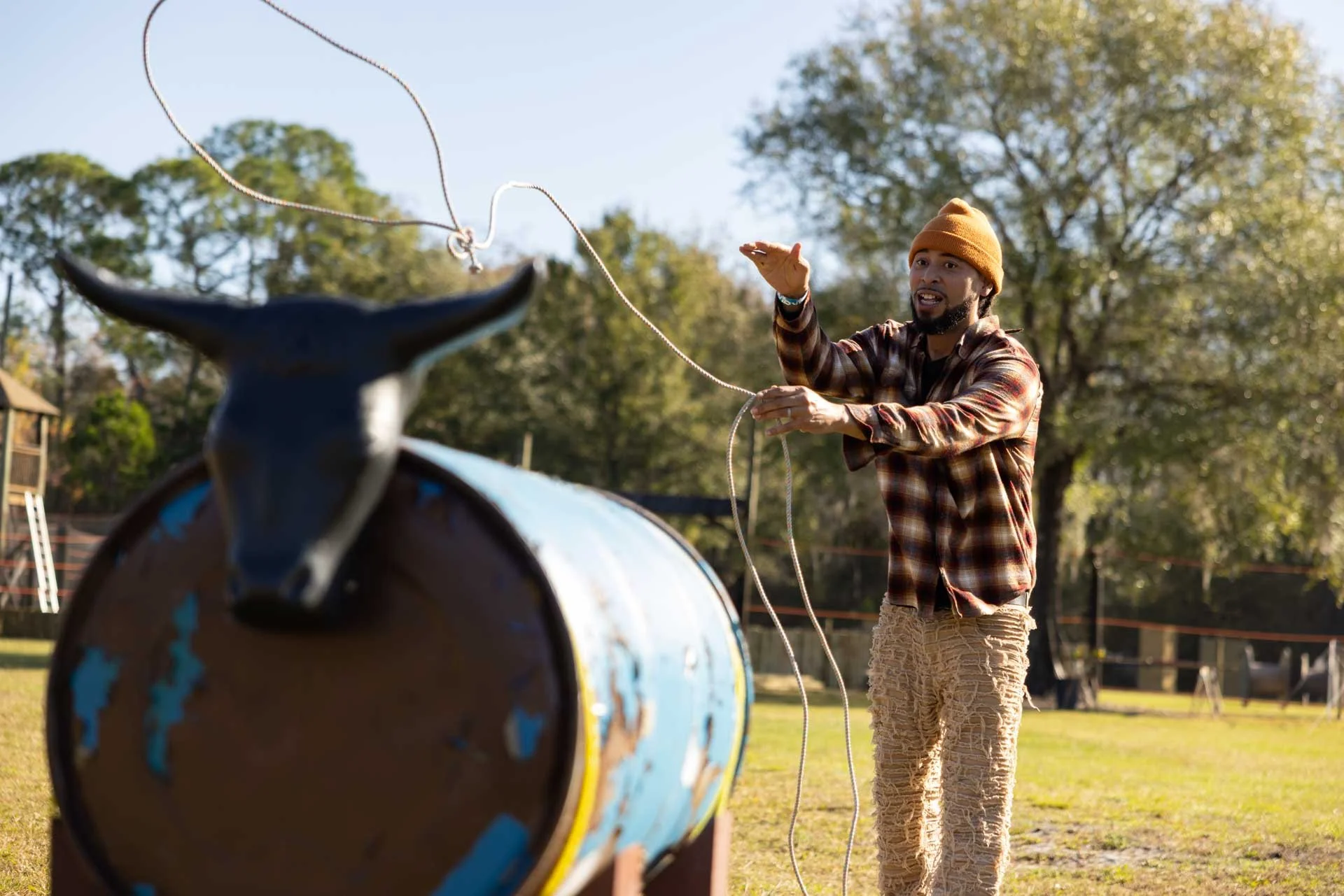 A man with a brown beanie and plaid shirt is playing a drum with a stick outdoors in a park with trees.