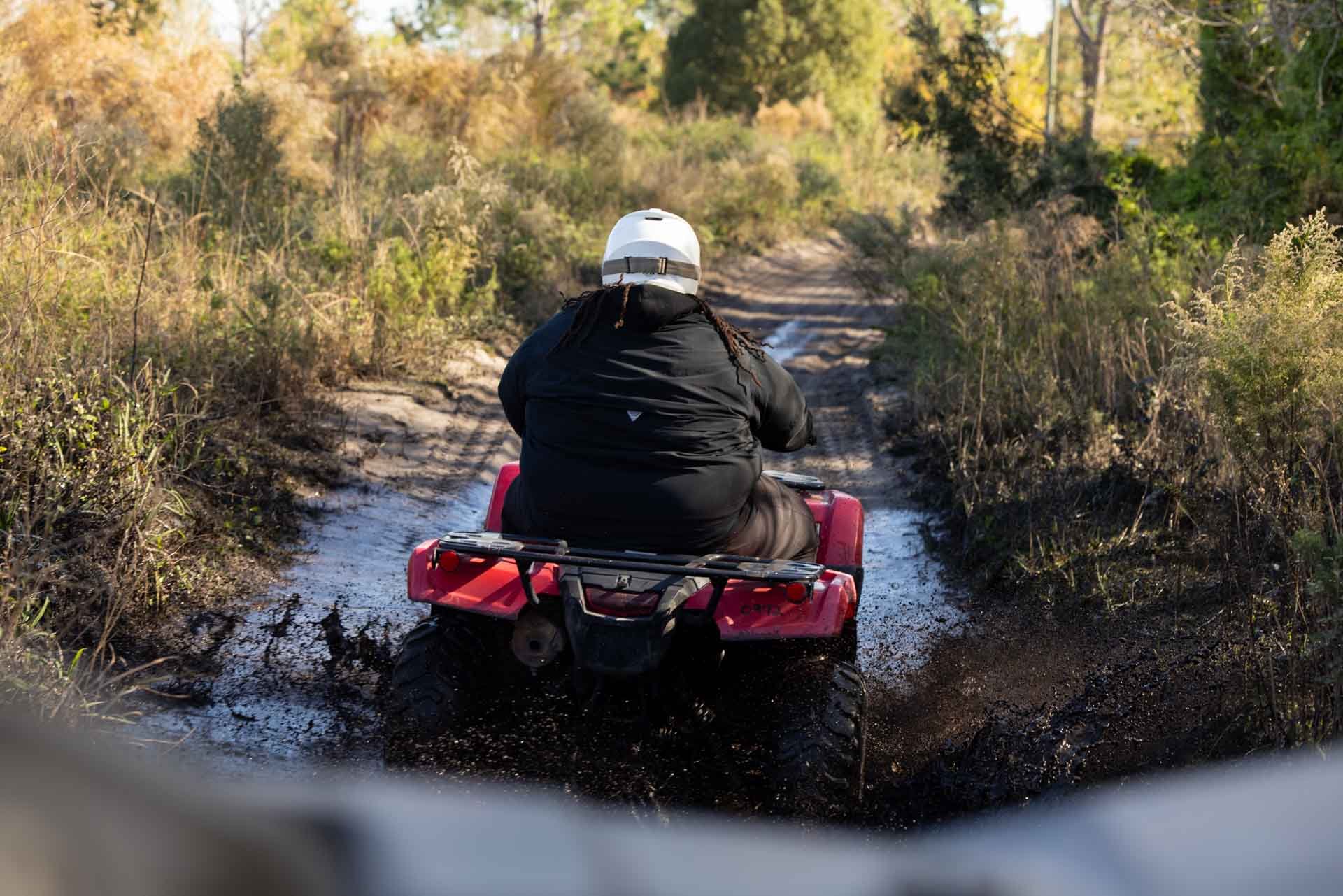 Person riding an ATV on a muddy trail through a wooded area, viewed from behind.