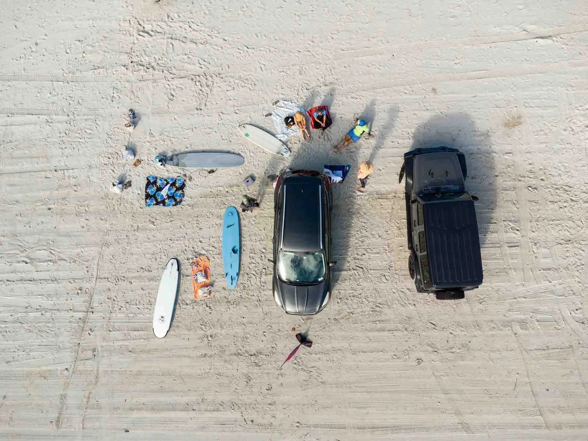 Aerial view of a beach with two parked cars, surfboards, and people preparing for surfing.