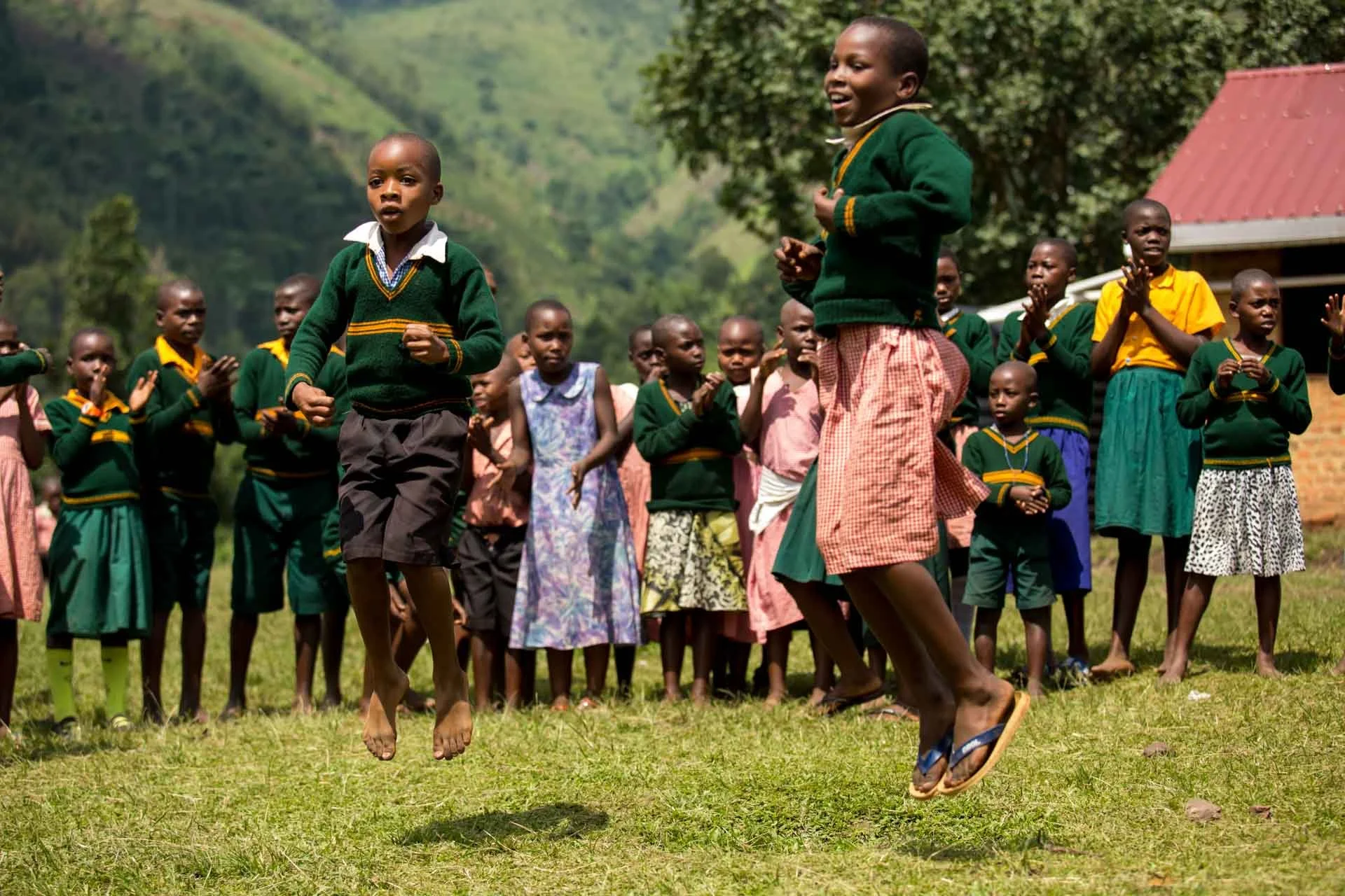 Children playing and jumping outdoors in a grassy area, with others watching, surrounded by green trees and buildings.