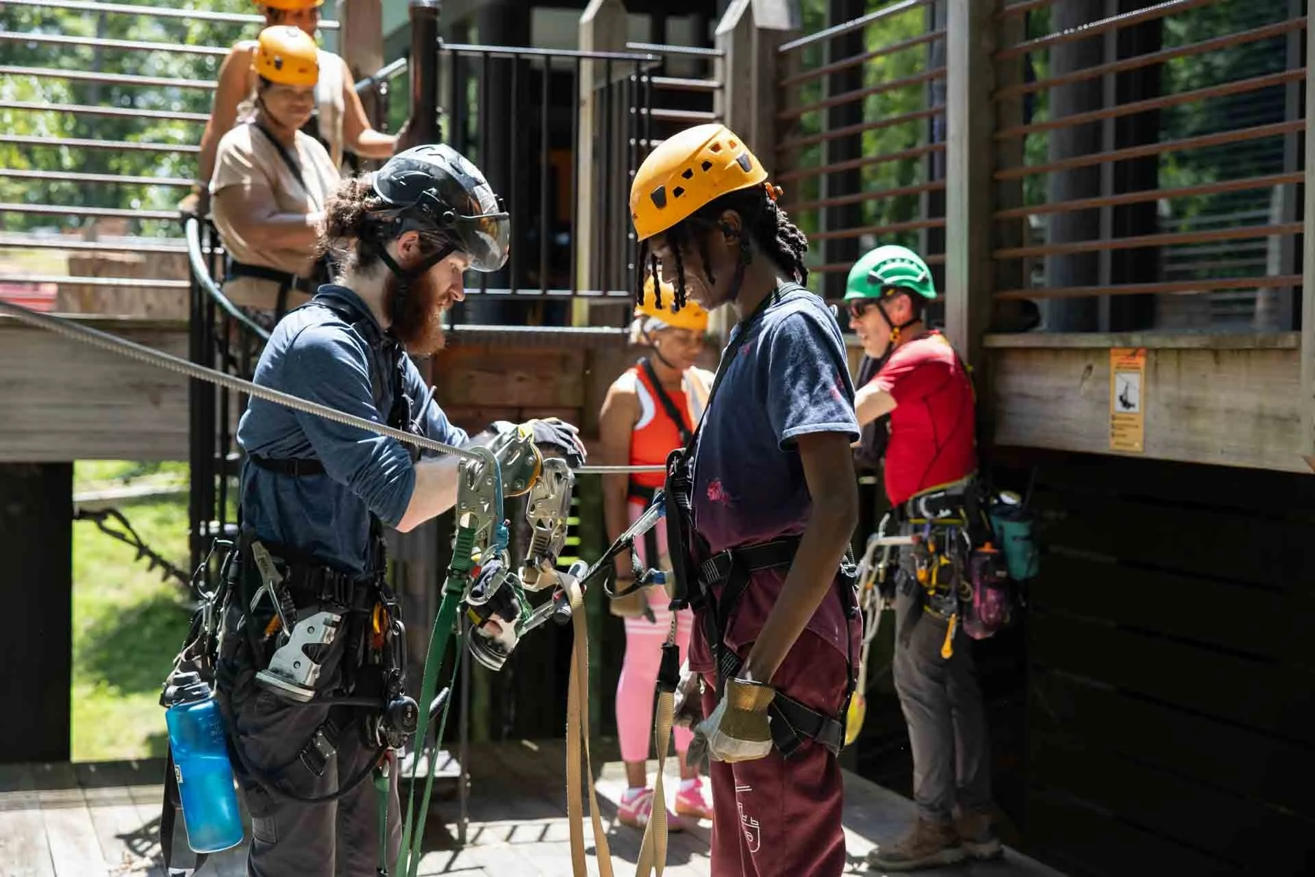 Group of people at a treetop adventure park, wearing helmets and harnesses, preparing for a zip line or climbing activity.