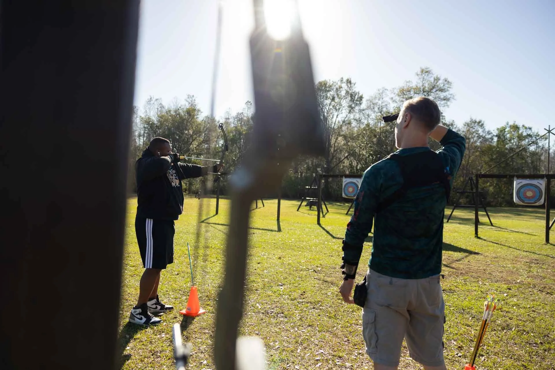 Two young men practicing archery outdoors on a sunny day, with target archery targets set up in the background.