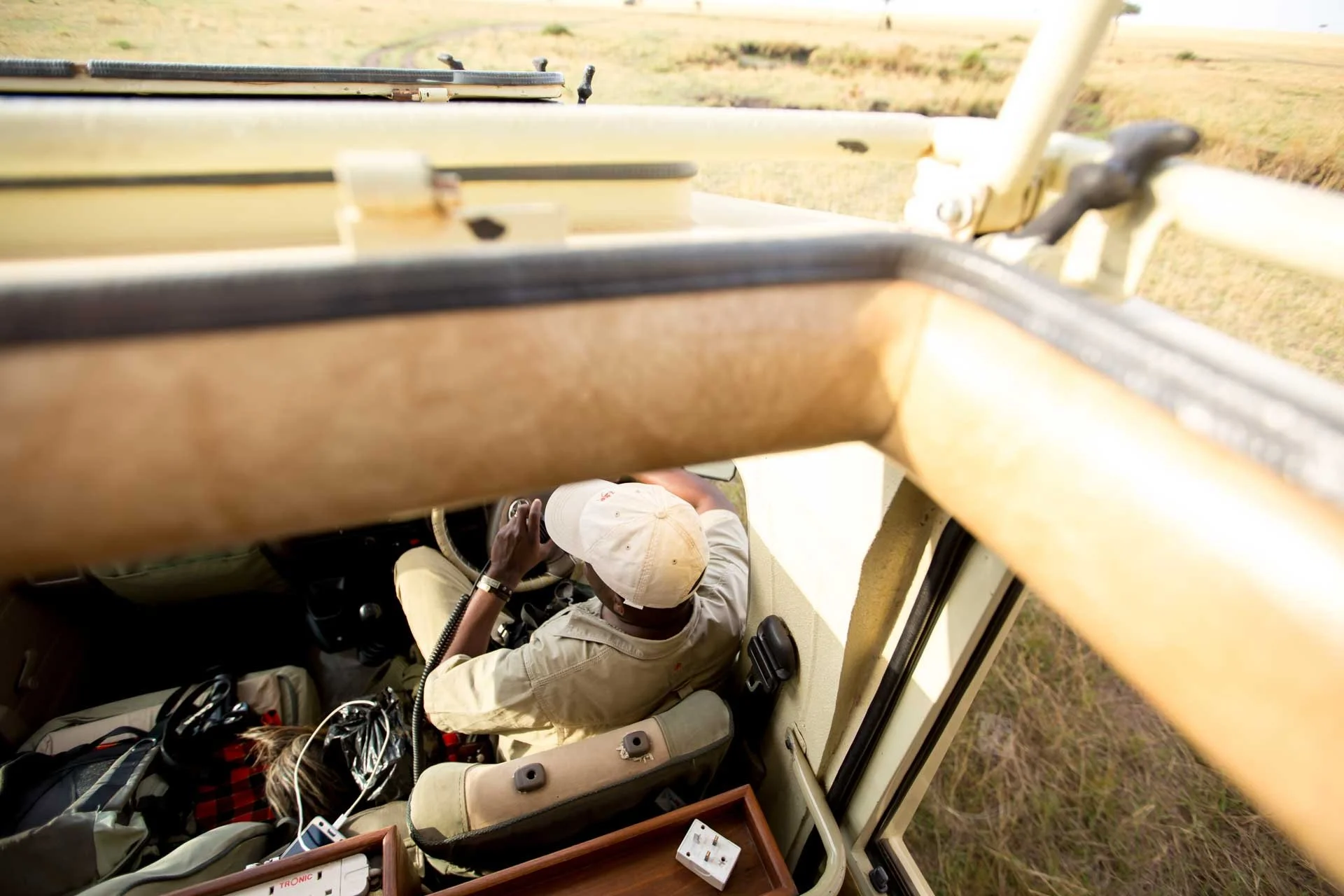 A person sitting in the driver's seat of a vehicle, looking out the window, with a field visible outside.