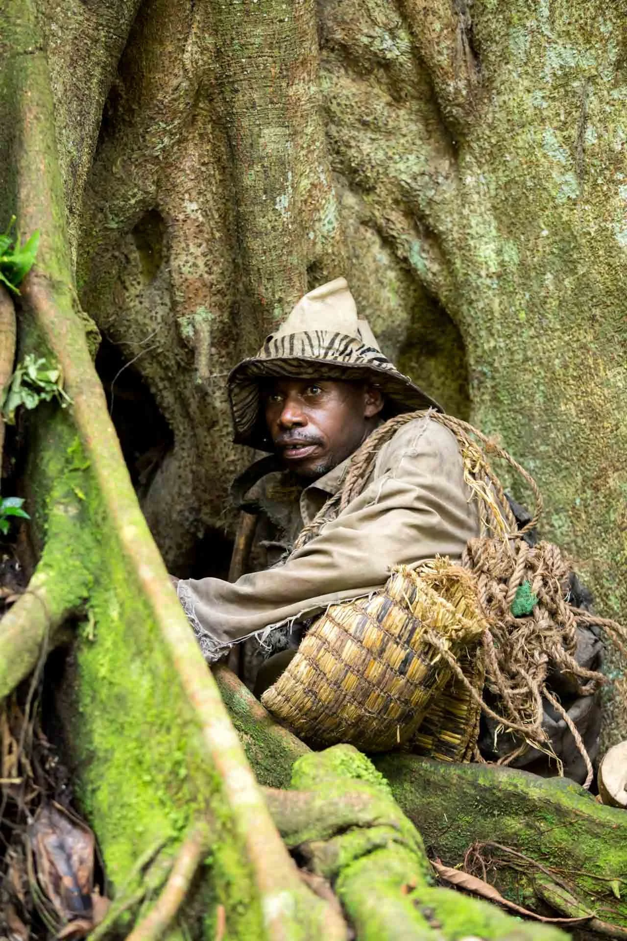 A man wearing a camouflage hat and tan jacket with a woven basket and ropes, sitting amid large tree roots with moss and greenery, looking alert.