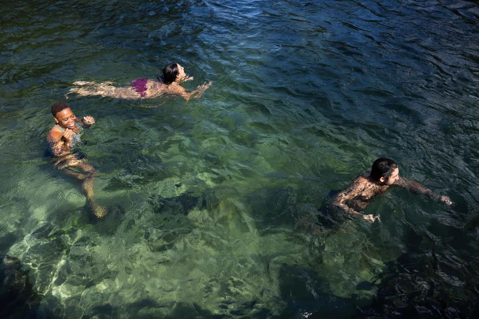 Three people swimming in clear water with visible rocks, a woman in purple swimsuit, a woman in green swimsuit, and a man with dark hair and beard.