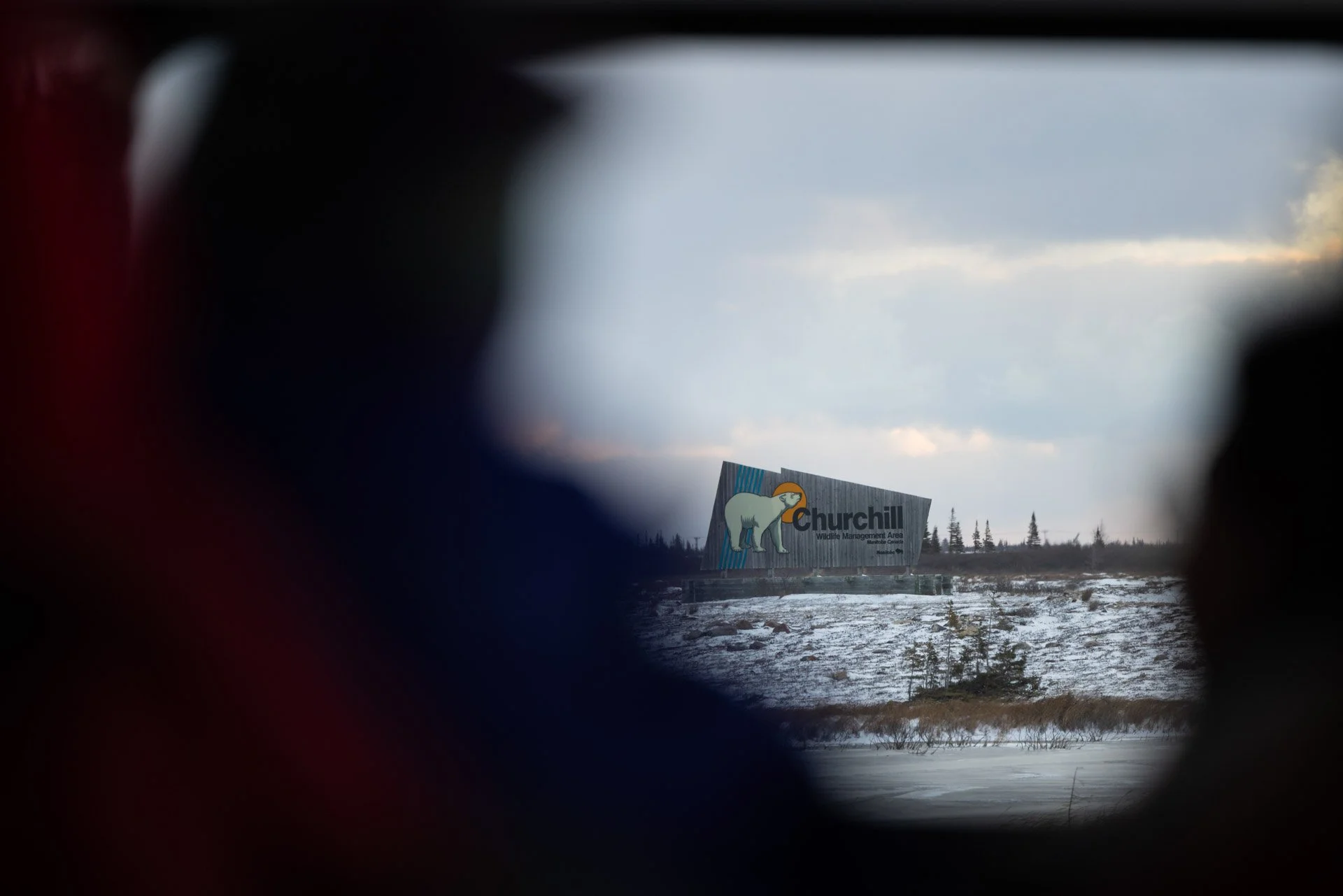 View through binoculars showing a sign for Churchill Wildlife Management Area with a polar bear illustration, on a snowy landscape with a cloudy sky.