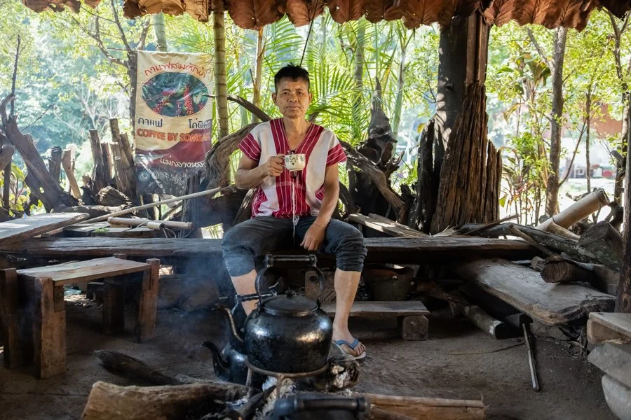 A man sitting inside a rustic open-air structure, holding a cup, with a teapot on the fire in front of him, surrounded by wooden logs and greenery.
