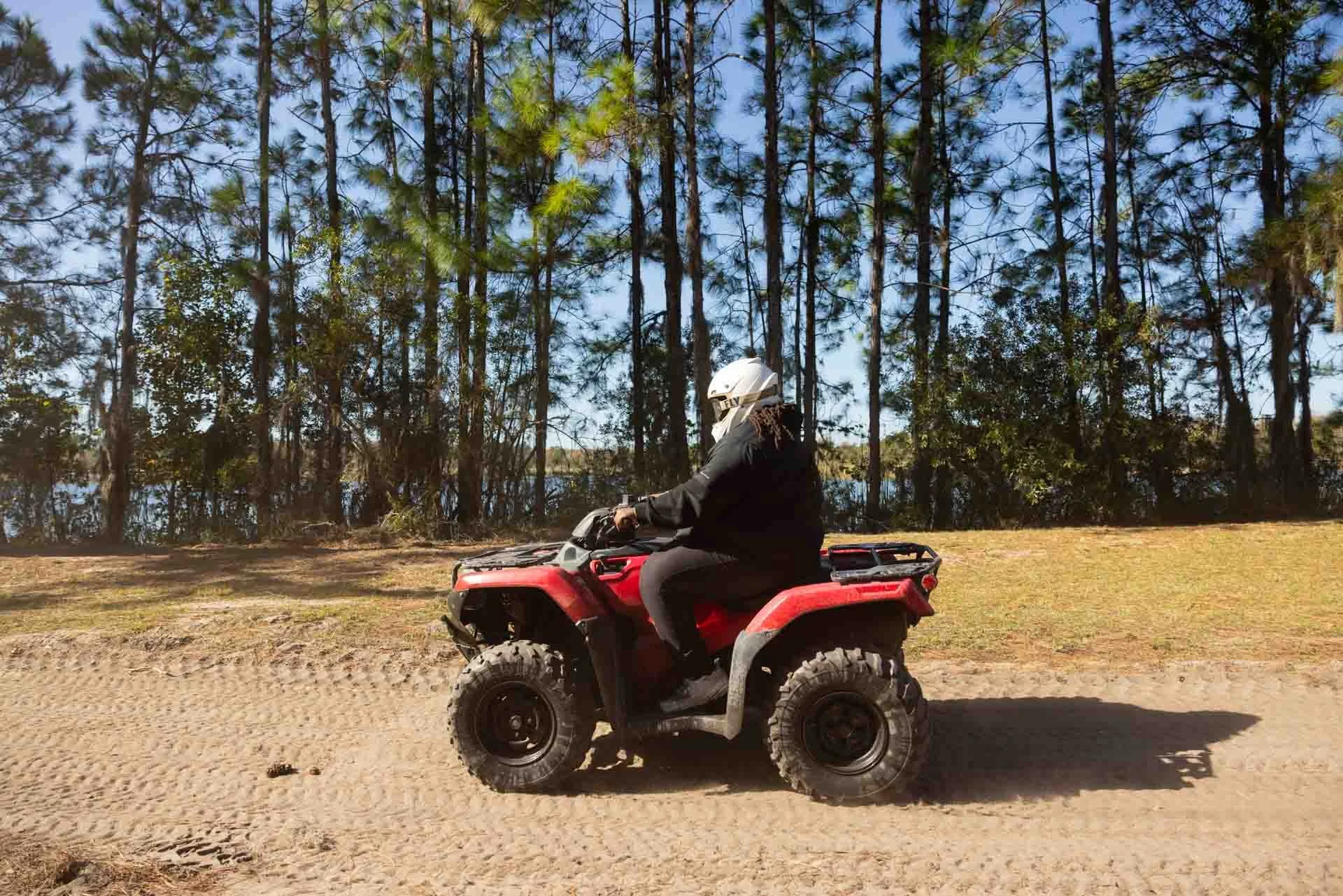 A person wearing a helmet and black clothing riding a red all-terrain vehicle on a dirt path near a wooded area.
