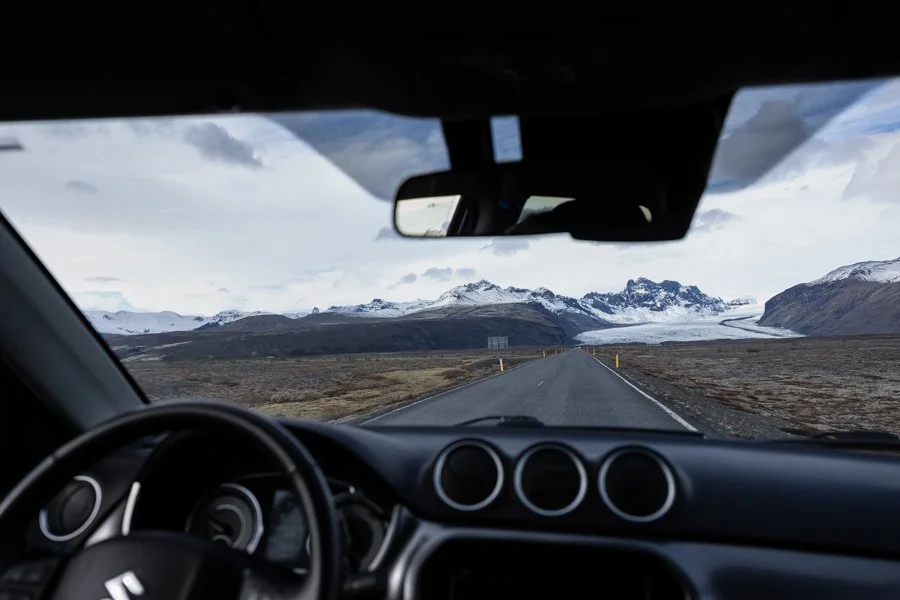 View from inside a car showing a road leading to snow-capped mountains under cloudy skies.