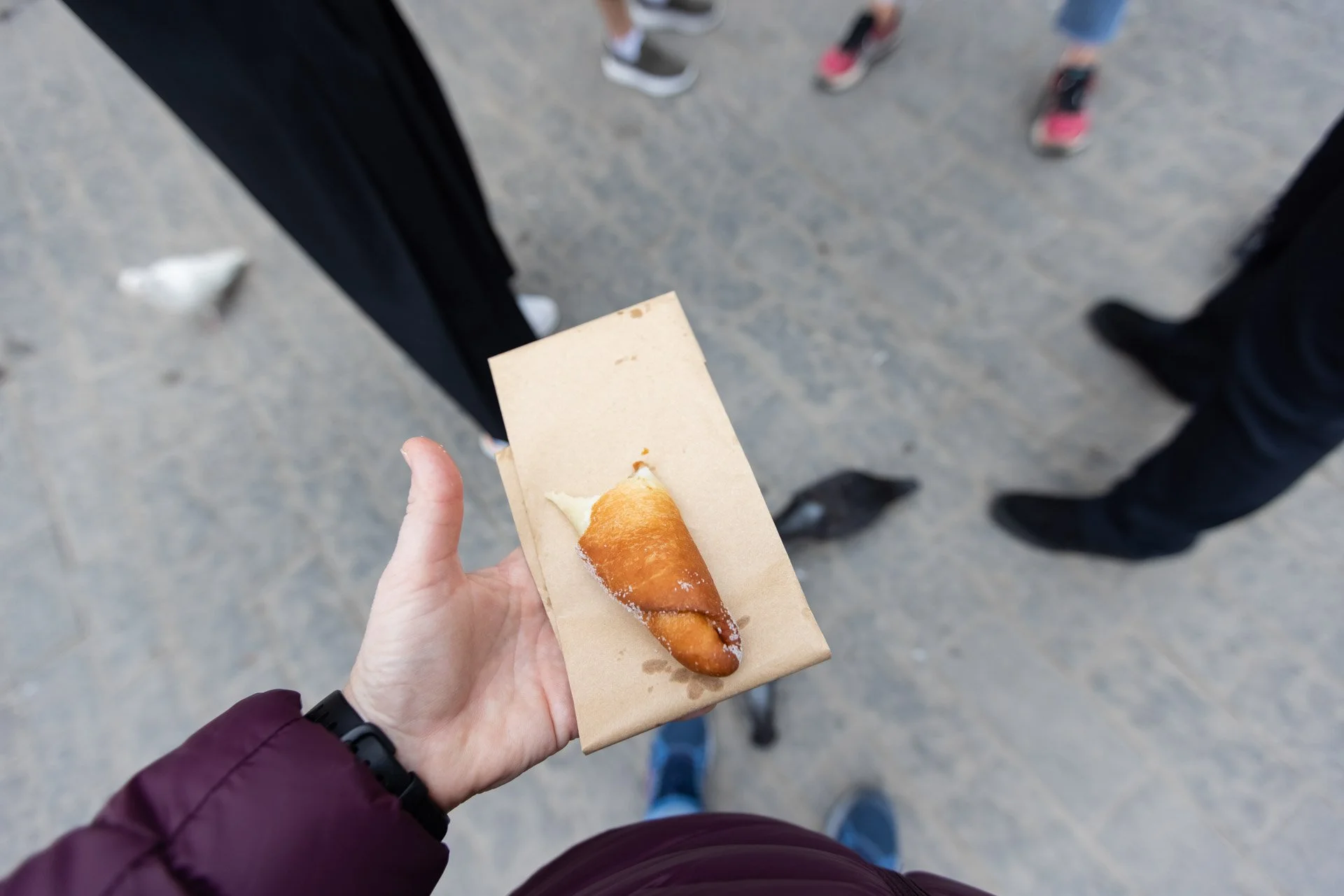 Person holding a paper bag with a cone of ice cream or frozen treat, surrounded by people whose legs are visible on a paved surface.