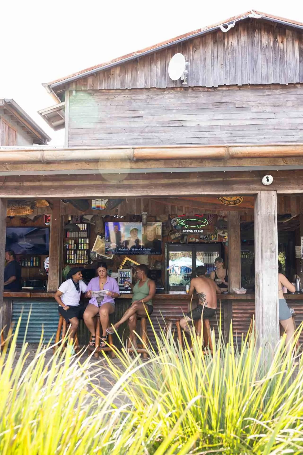 People sitting at a bar on a patio with wooden beams, neon signs, and a television, in a casual outdoor setting.
