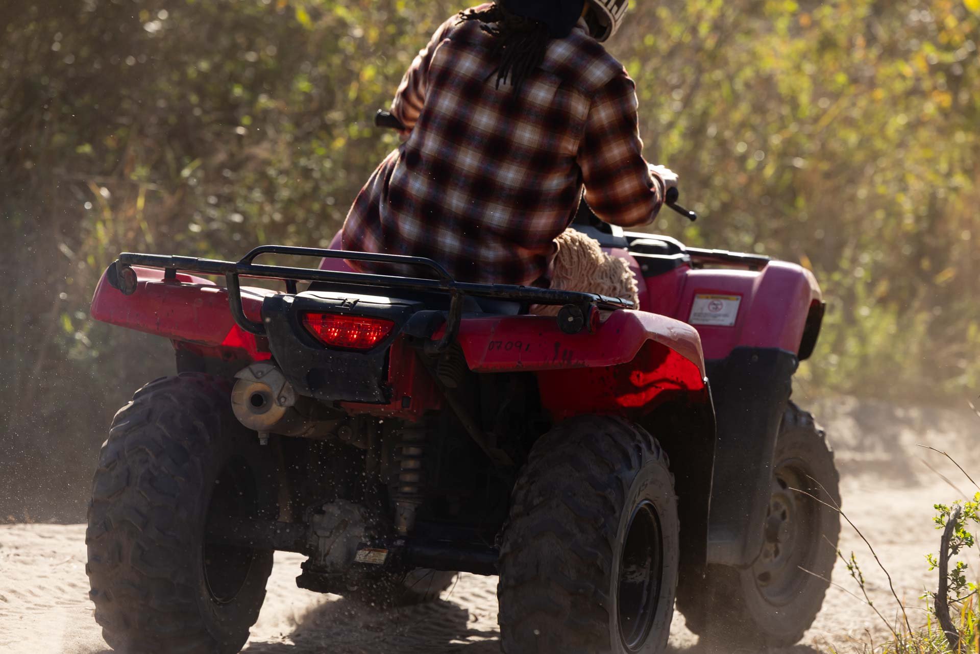 A person wearing a plaid shirt riding a red ATV on a dirt trail in a wooded area.