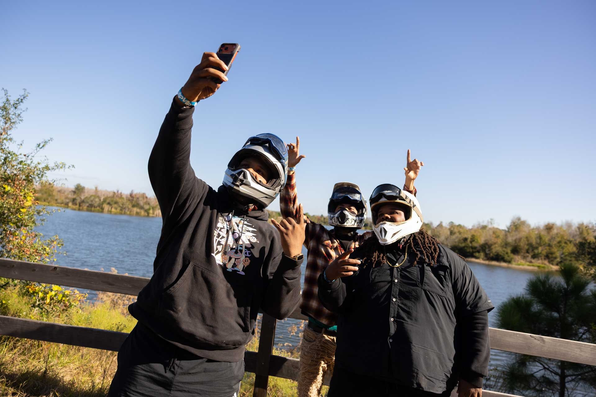 Three people wearing motorcycle helmets taking a selfie outdoors near a lake with trees in the background.