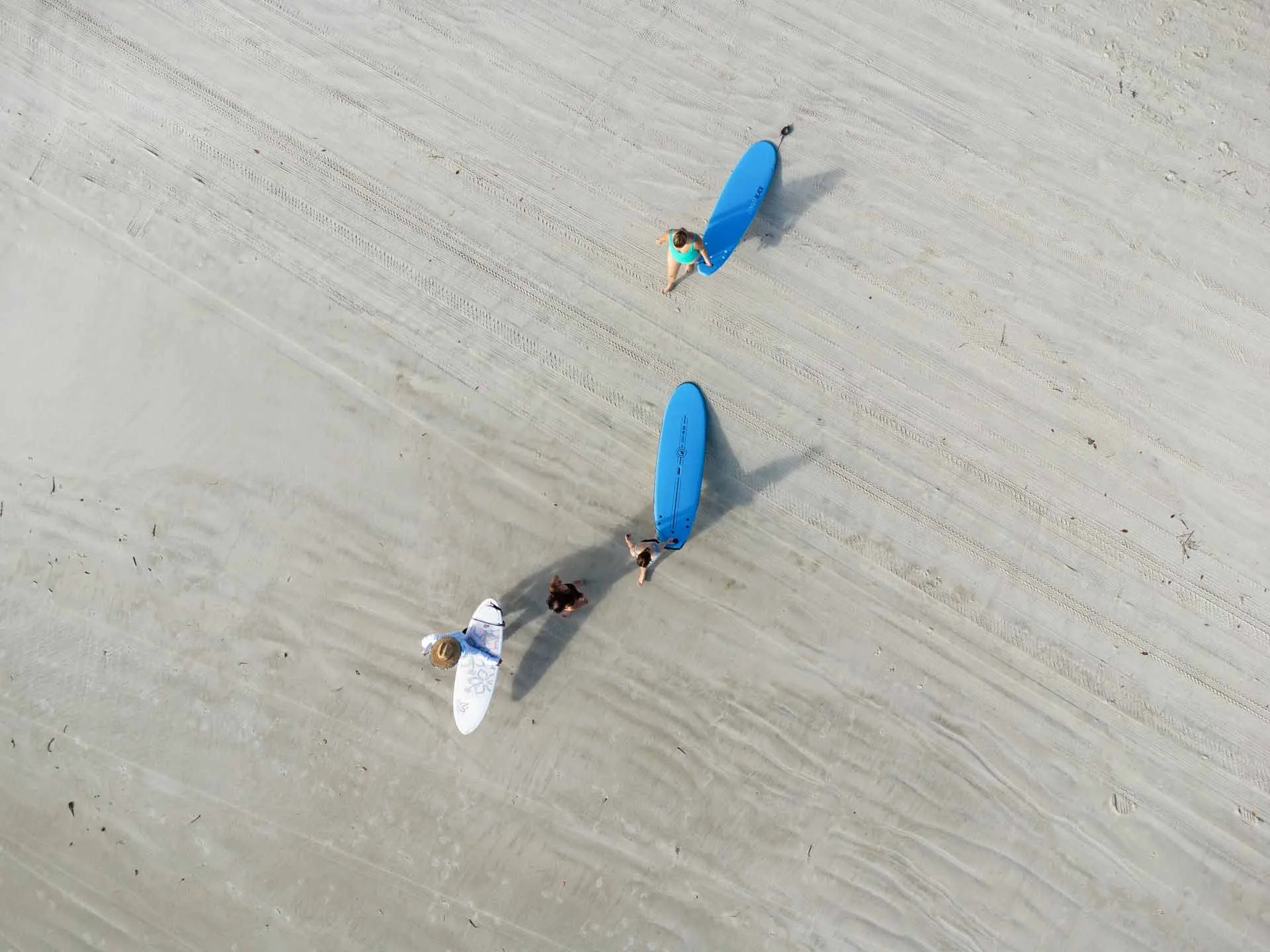 Three people on a sandy beach with surfboards, one on a white surfboard, and two on blue surfboards, seen from above.