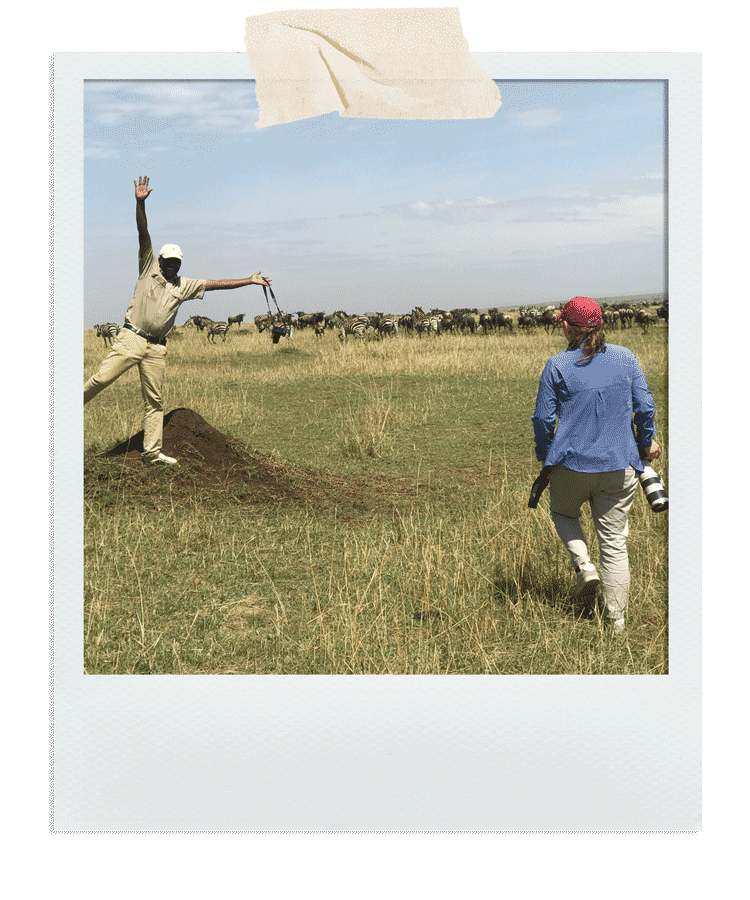 A person standing on a mound of dirt in a grassy plain with zebras in the background, waving to the camera. Another person, holding a camera, walks towards them. The sky is partly cloudy.