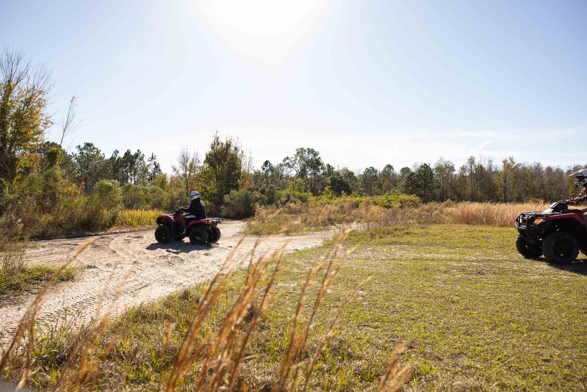 Two people riding all-terrain vehicles (ATVs) on a dirt path in a grassy, wooded area on a sunny day.