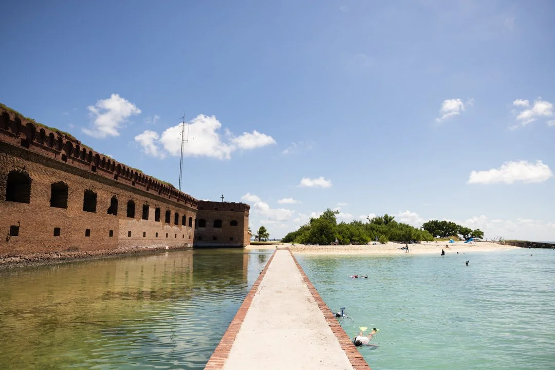 A historic brick fort along a narrow walkway extending into calm water, with a sandy beach and green trees in the background and a blue sky with scattered clouds.