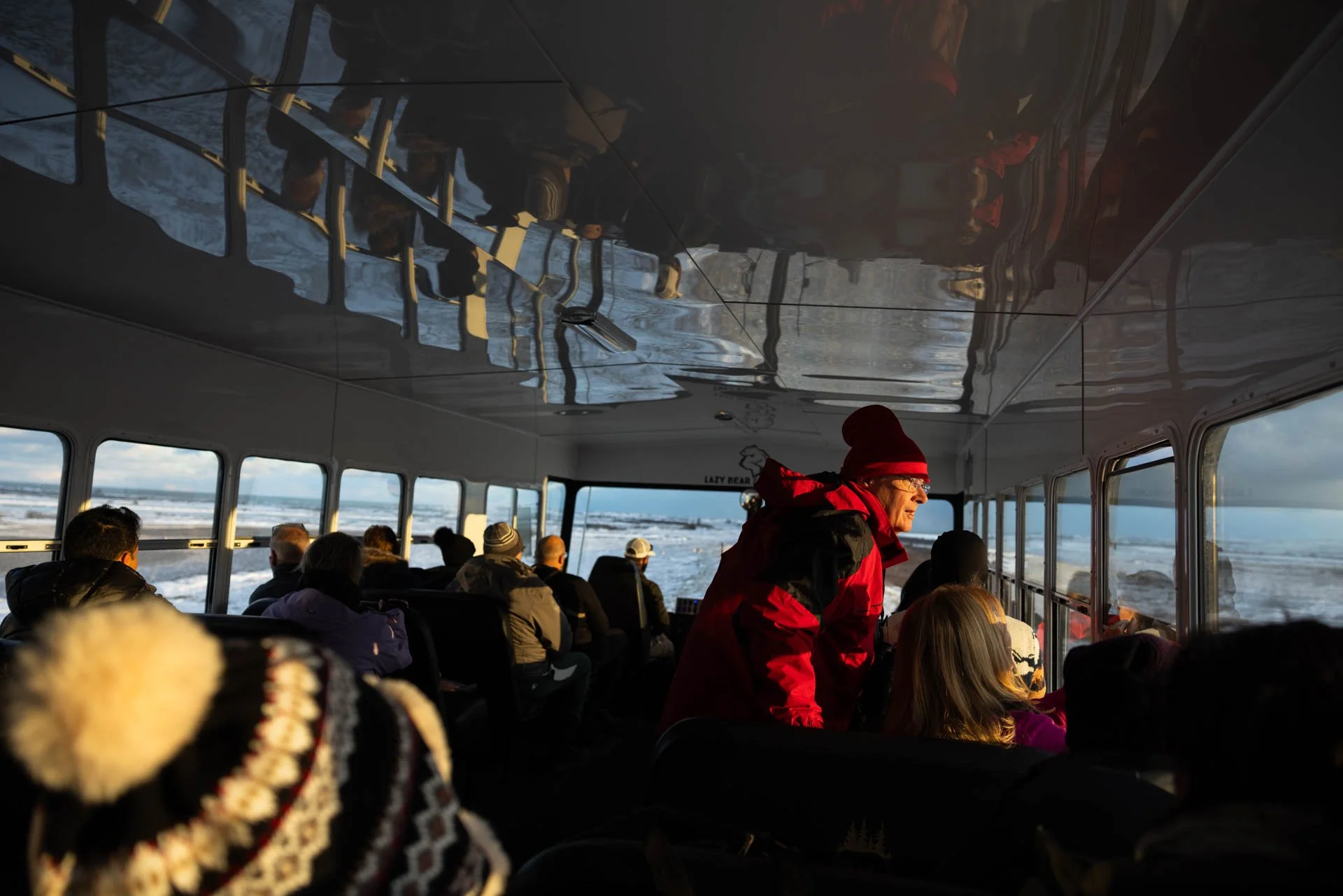 Interior of a bus or boat with passengers seated, looking out windows at snow-covered landscape, with one person standing in red winter coat and hat.