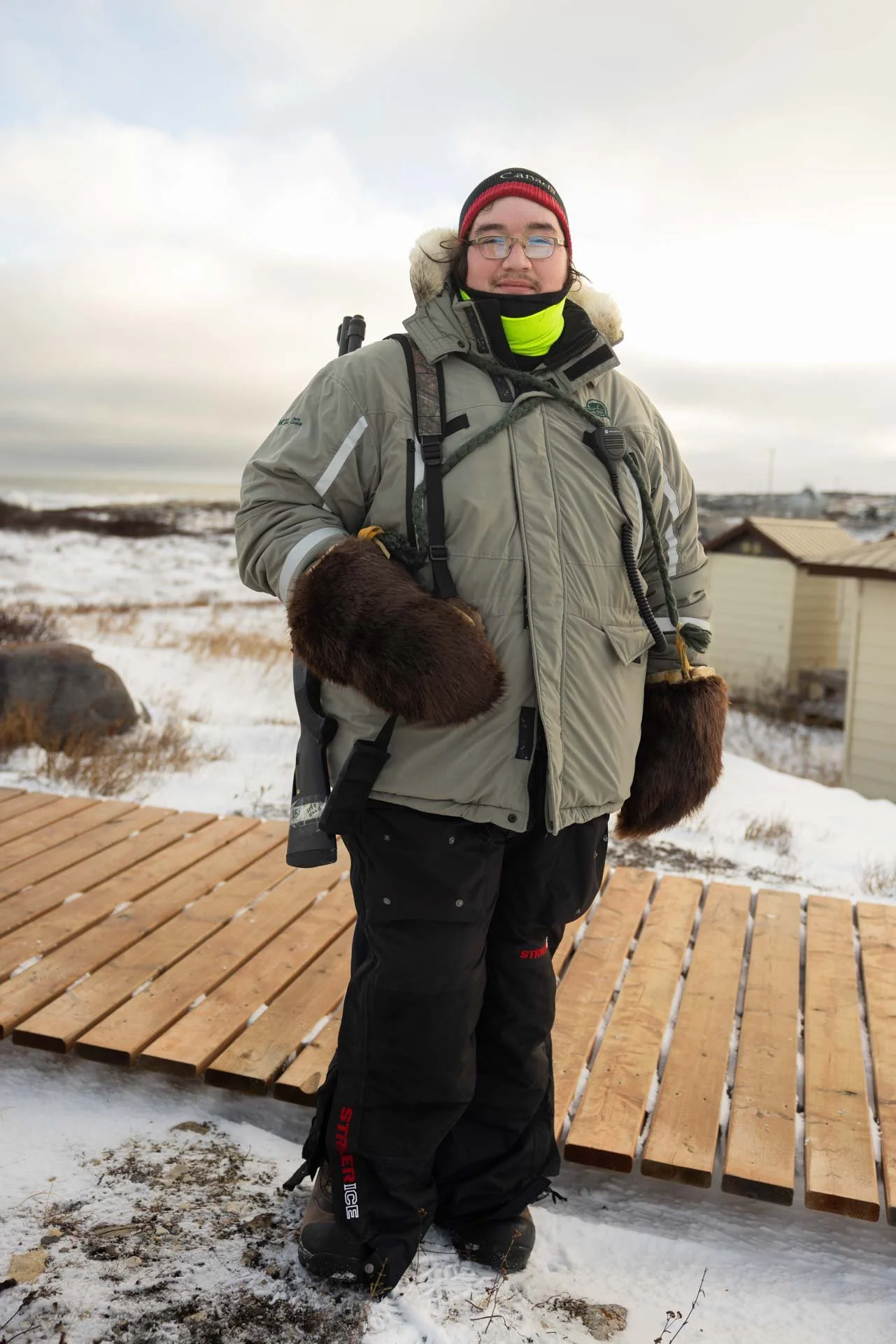 A person dressed in winter gear, including a gray jacket, black pants, and brown fur-lined mittens, standing on a snowy wooden deck in a snowy landscape with houses in the background.