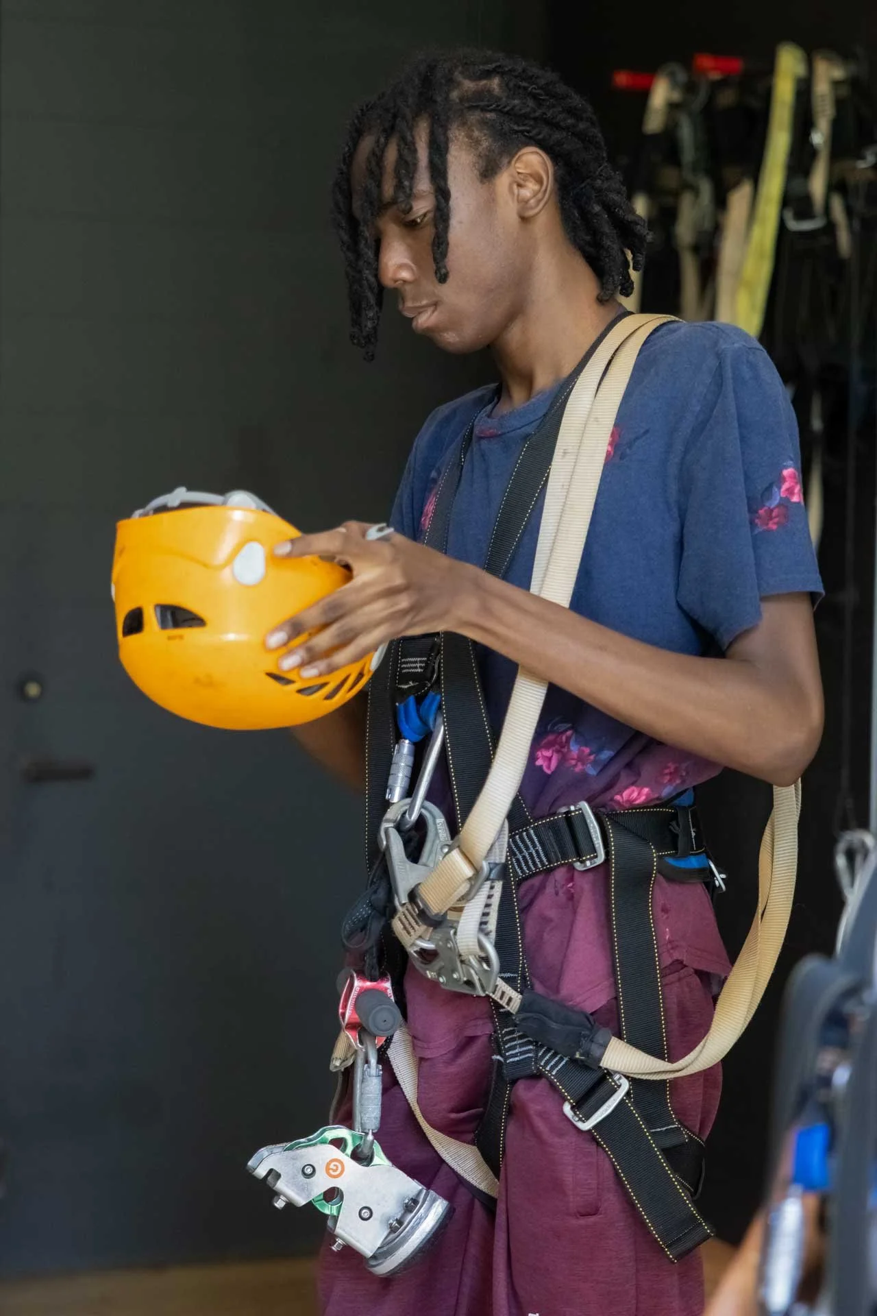 A person with dreadlocks wearing climbing gear holding a yellow helmet.