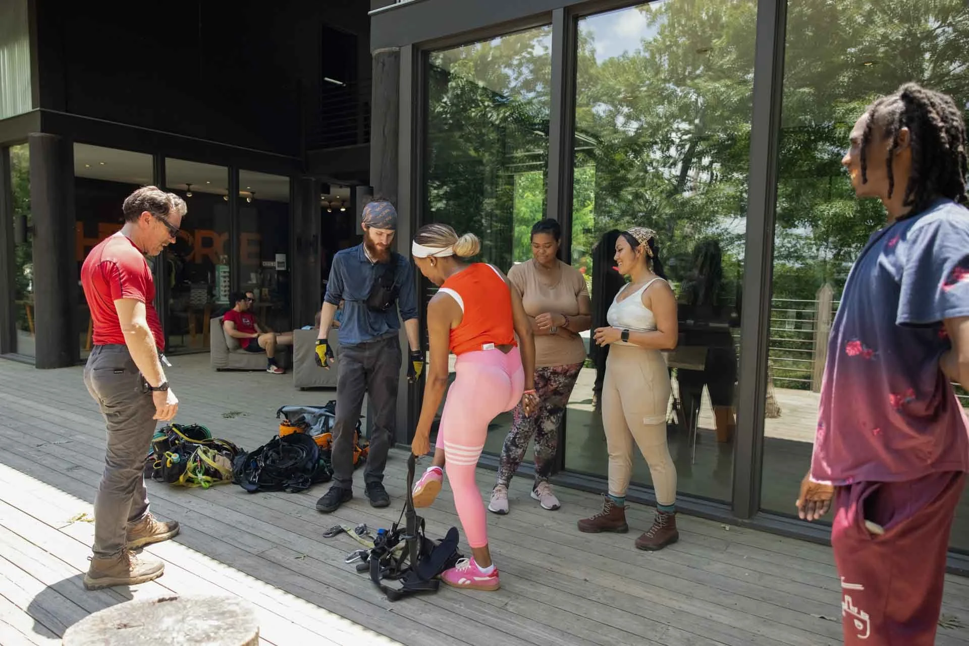 A group of six people gathered outside a building, preparing for an outdoor activity. One woman is putting on rollerblades while others watch or chat. There is equipment and backpacks on the ground nearby.