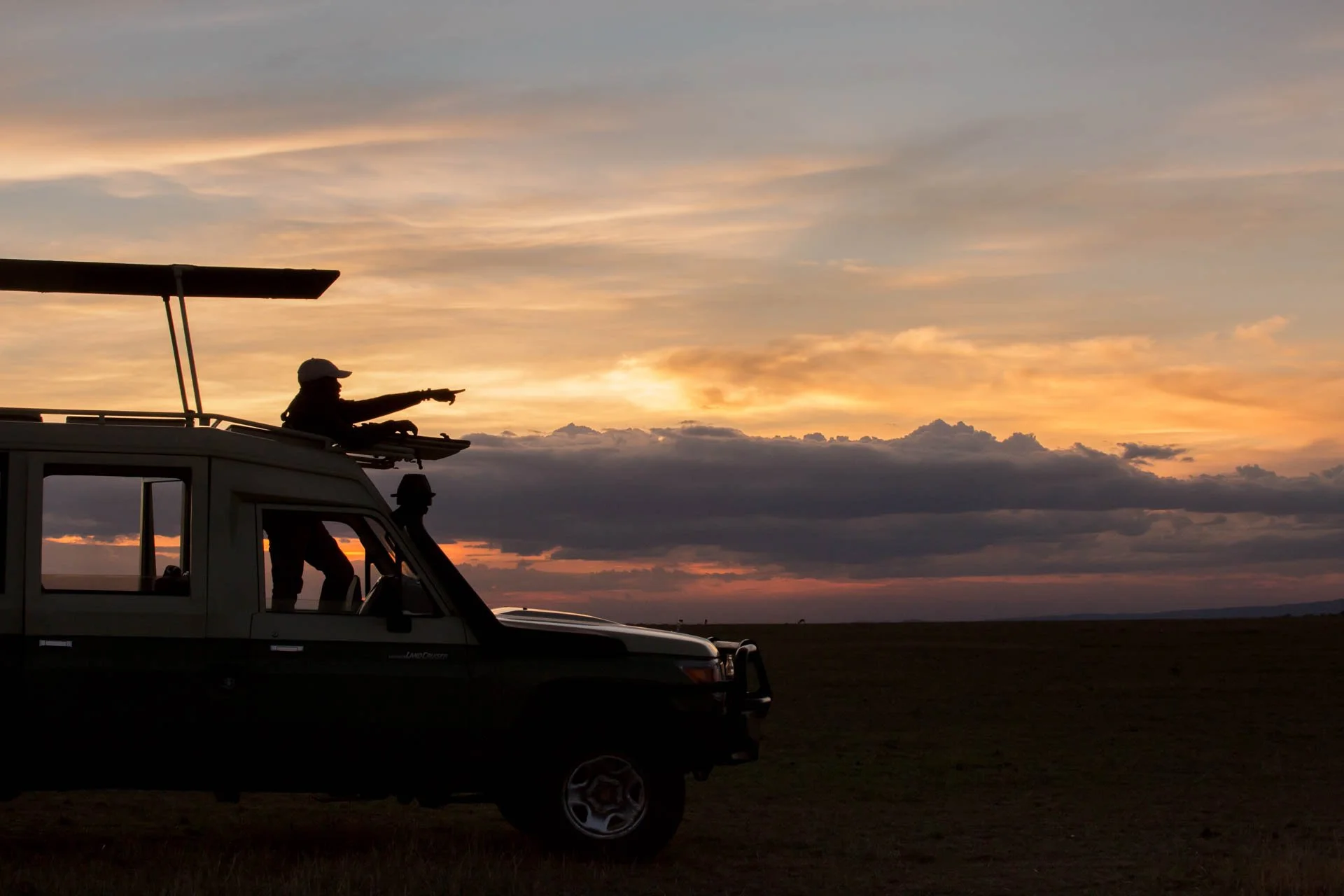 Silhouette of a safari vehicle with a guide pointing at the sunset sky in an open plain.