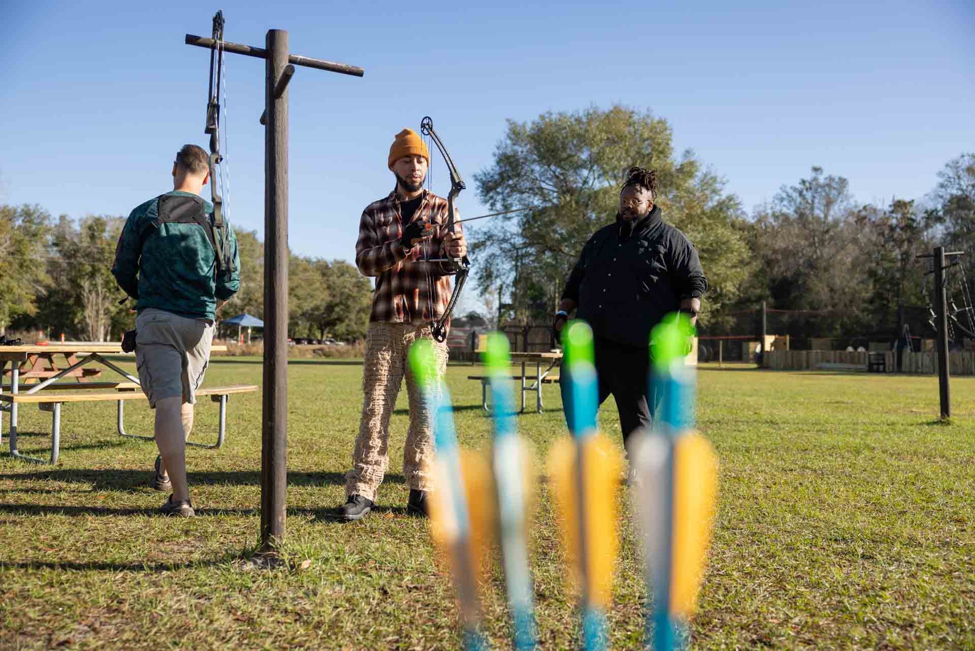 Three men are outdoors on a grassy field, one adjusting a bow, and the other two observing, with colorful lollipops in the foreground.