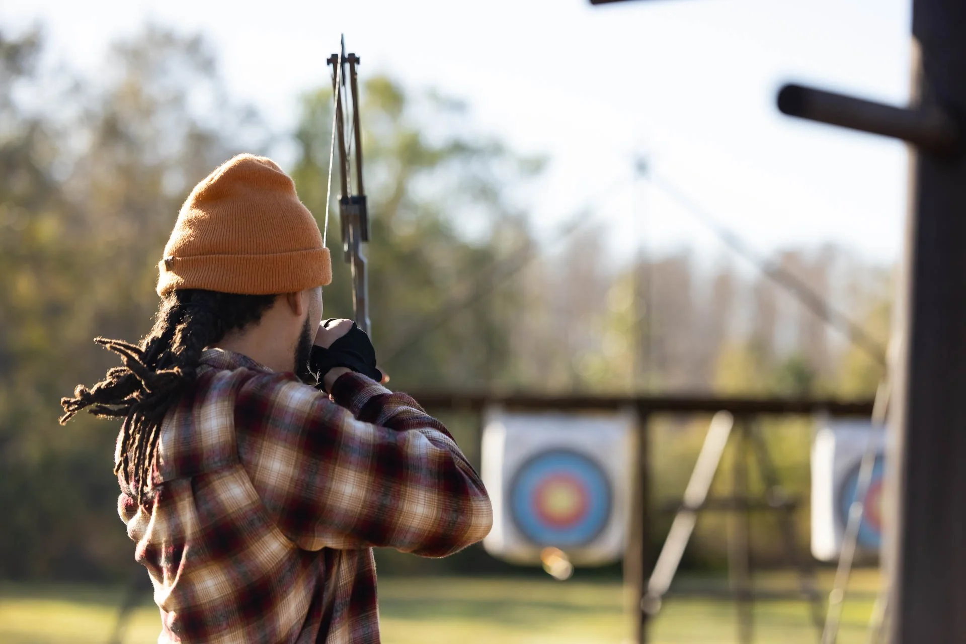 A man with dreadlocks wearing an orange beanie, plaid shirt, and gloves is shooting an arrow at an archery target outdoors during daytime.