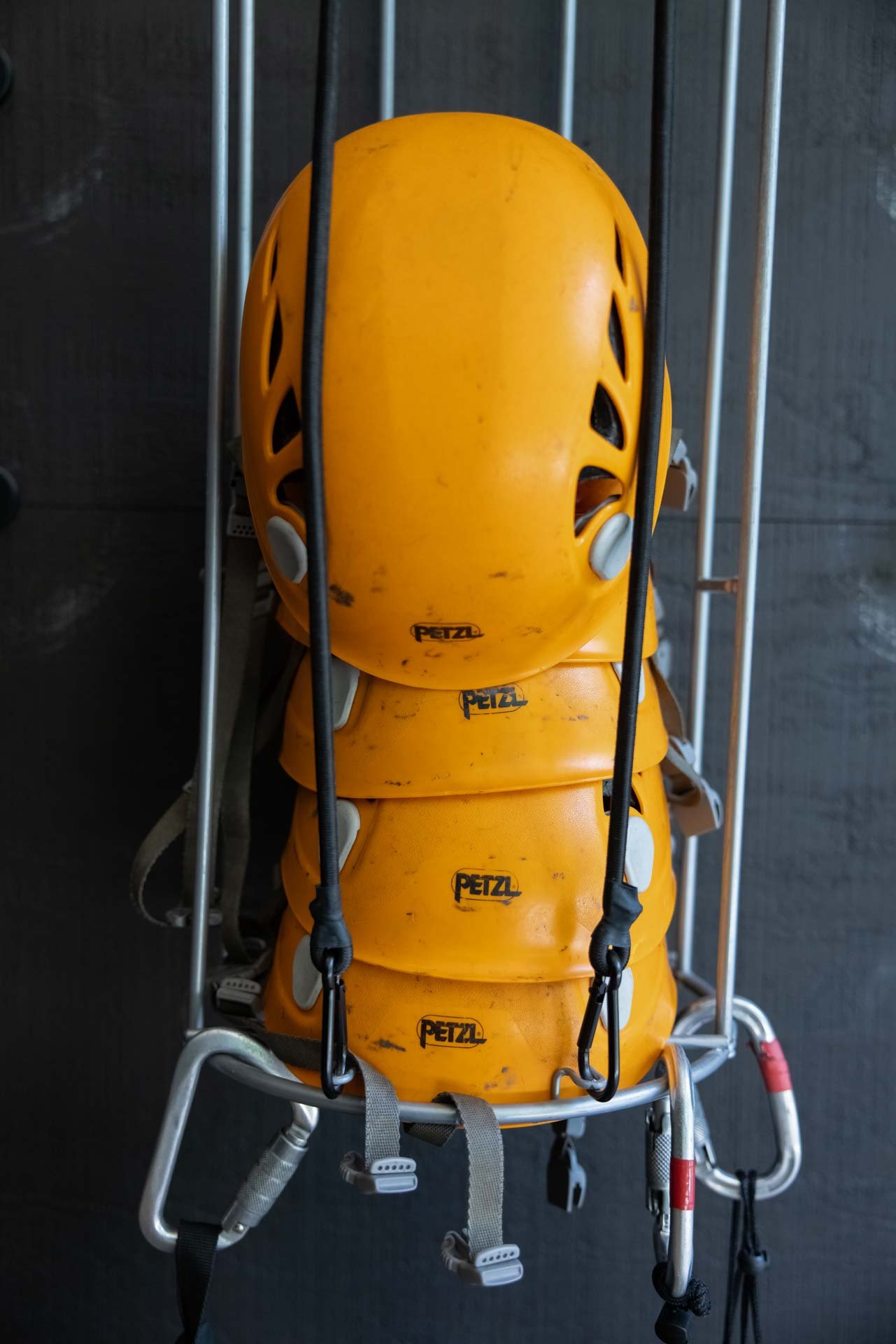Stack of four yellow Petzl climbing helmets with black logos, hanging on a metal rack with carabiners on the straps.