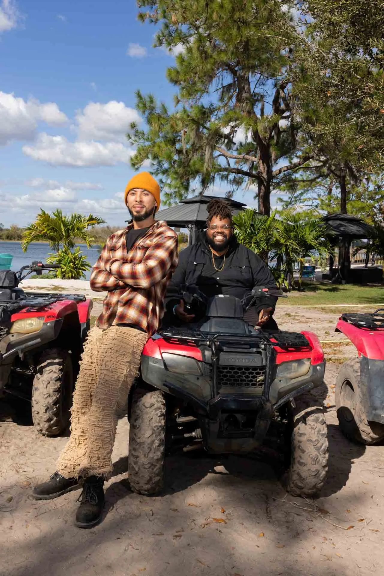 Two men with beards and black hair, one standing wearing an orange beanie and plaid shirt, the other sitting on a red ATV with black jacket, in an outdoor park with trees, shrubs, and a lake in the background.