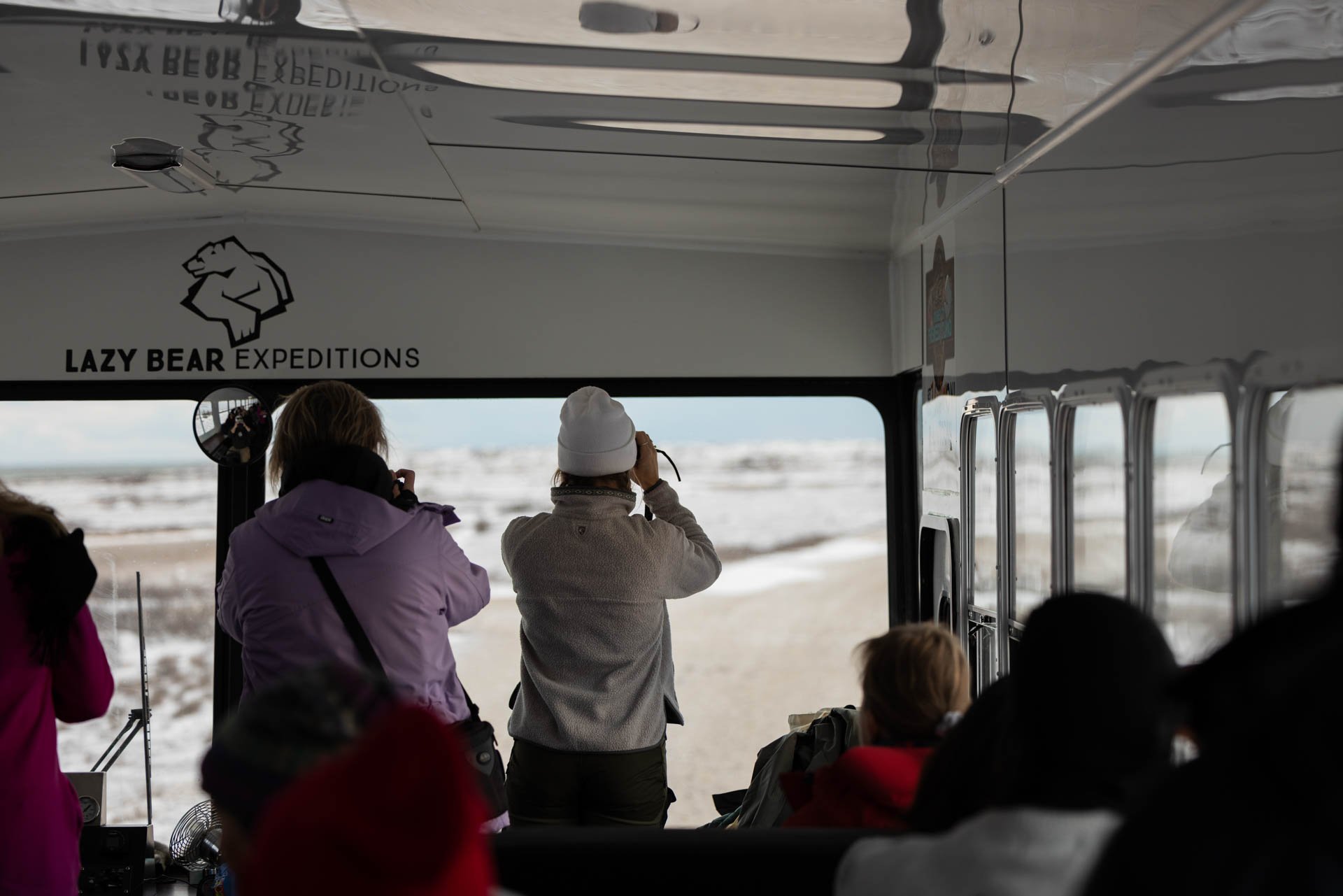 People on a bus or vehicle observing a snowy landscape outside, with some taking photos, under a 'Lazy Bear Expeditions' sign.