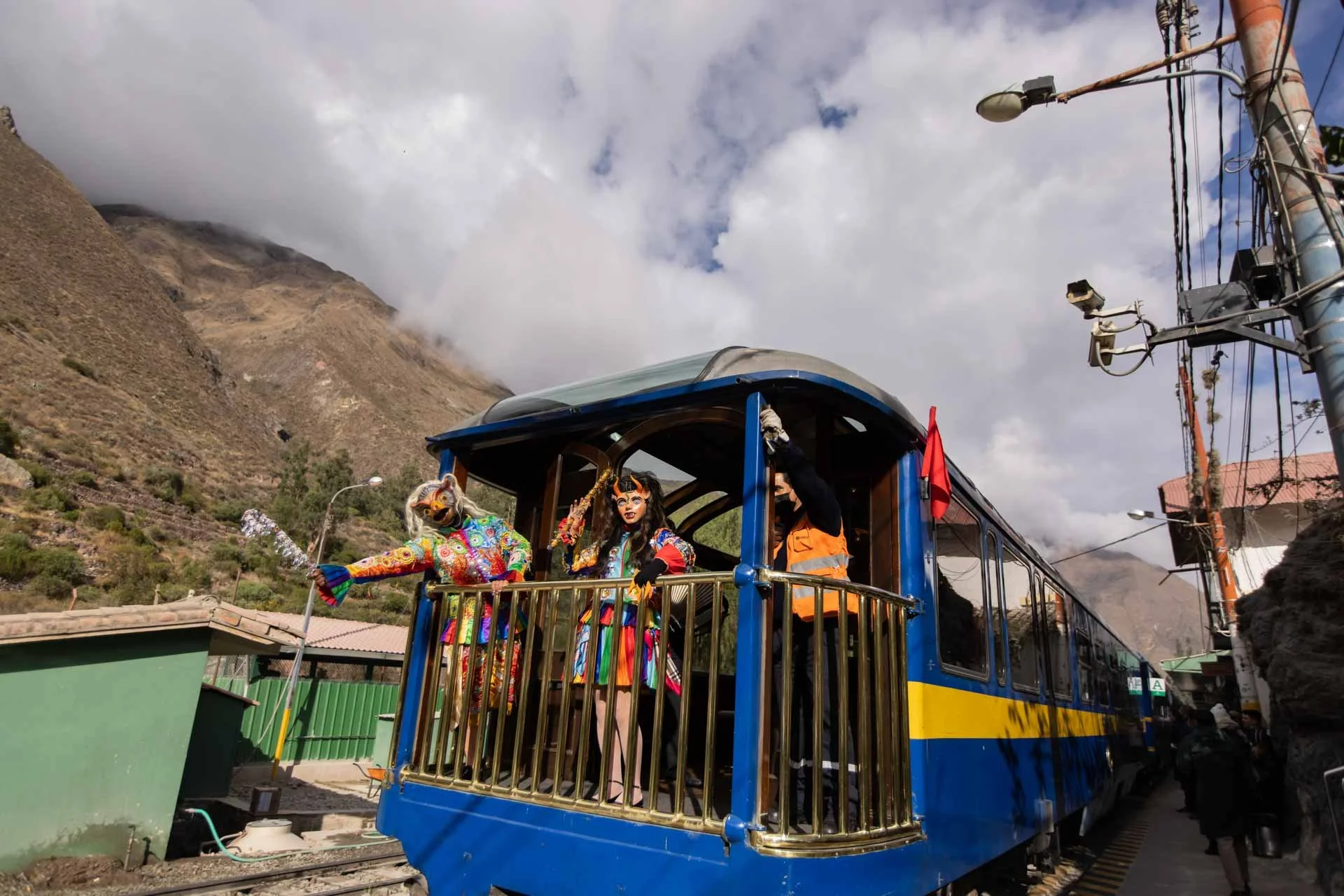 Colorful traditional dancers posing on a blue train carriage with mountains and cloudy sky in the background.