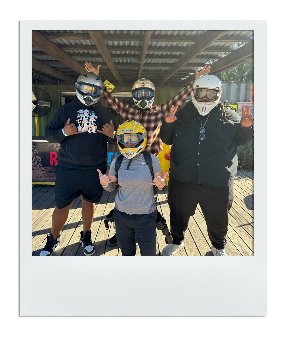 Five people wearing helmets and casual clothing, posing with hands raised or making gestures, outdoors on a wooden deck under a roof, with a fan, yellow sign, and other objects in the background.